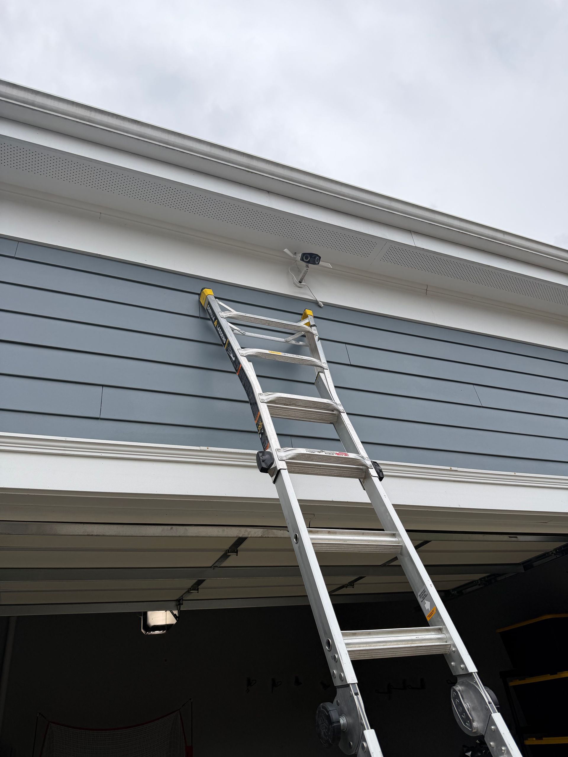 An aluminum extension ladder leans against a blue-sided house with white soffits and a garage door opening.