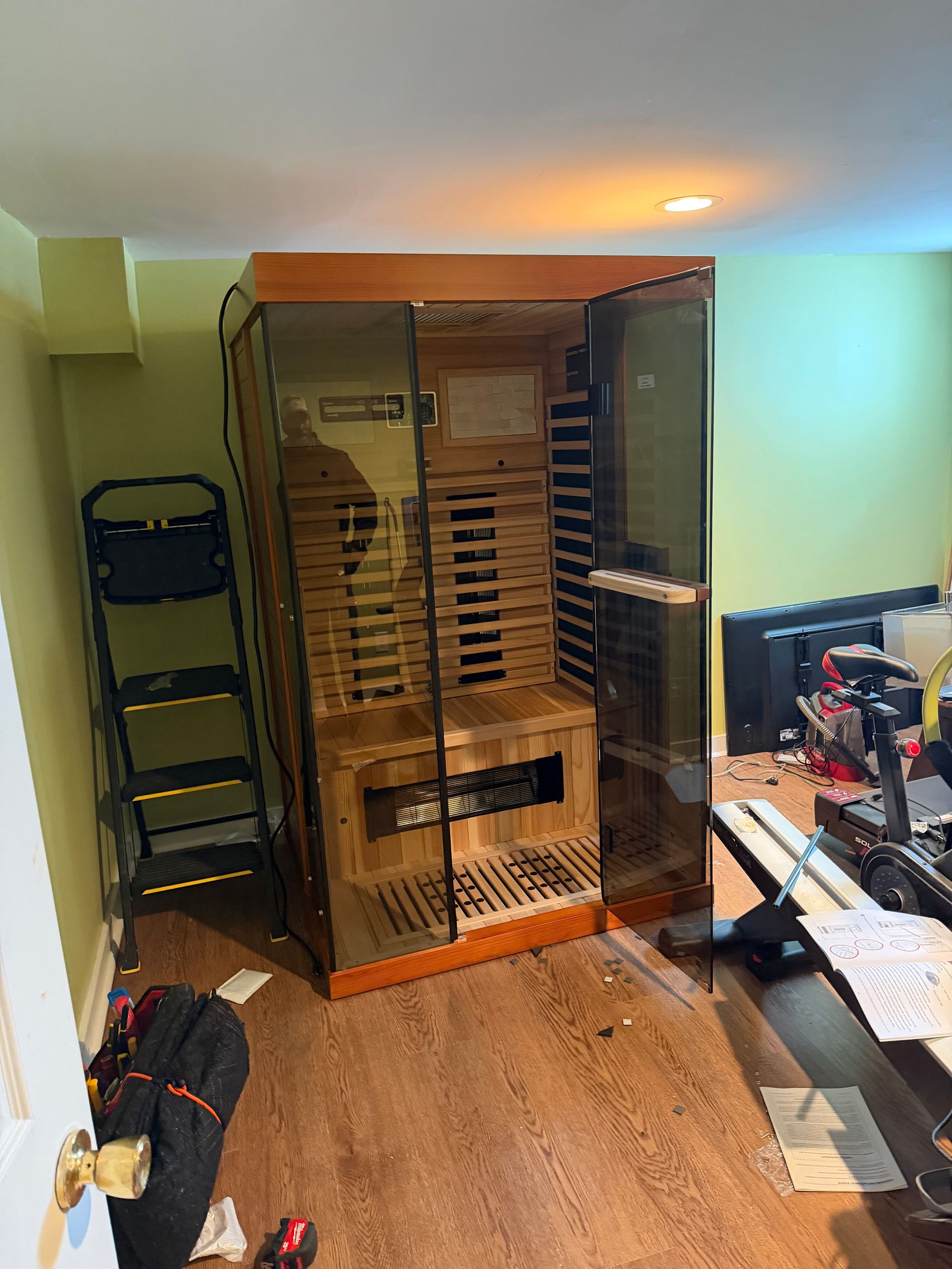 A wooden indoor infrared sauna stands in a room with light green walls, near a step ladder and various items on the floor.