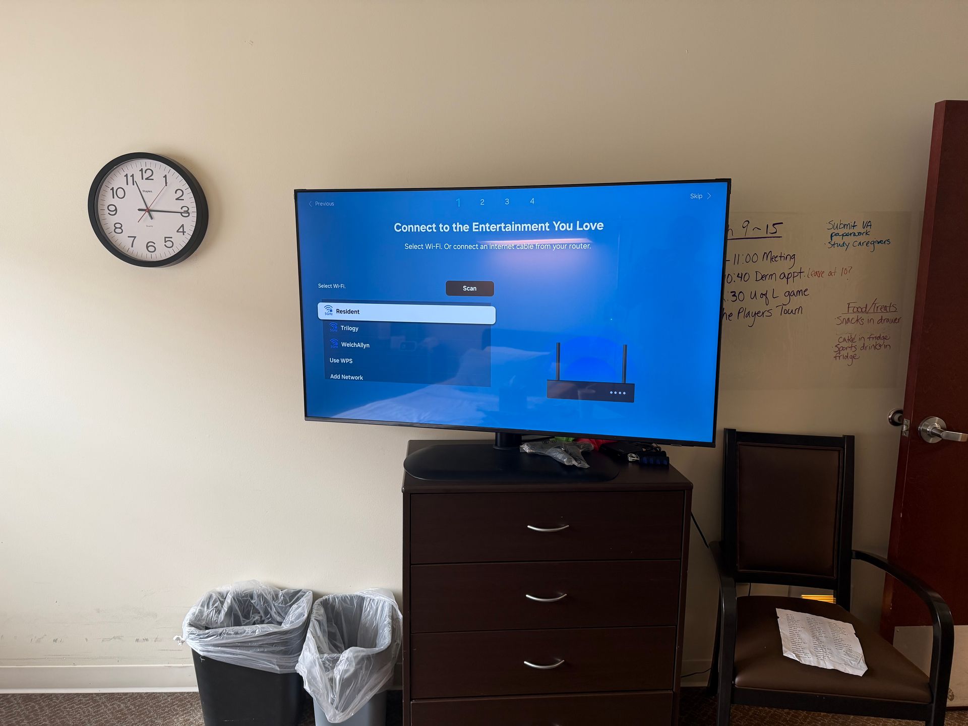 A TV on a wooden dresser displaying a blue setup menu, next to a wall clock, two trash cans, and a chair.