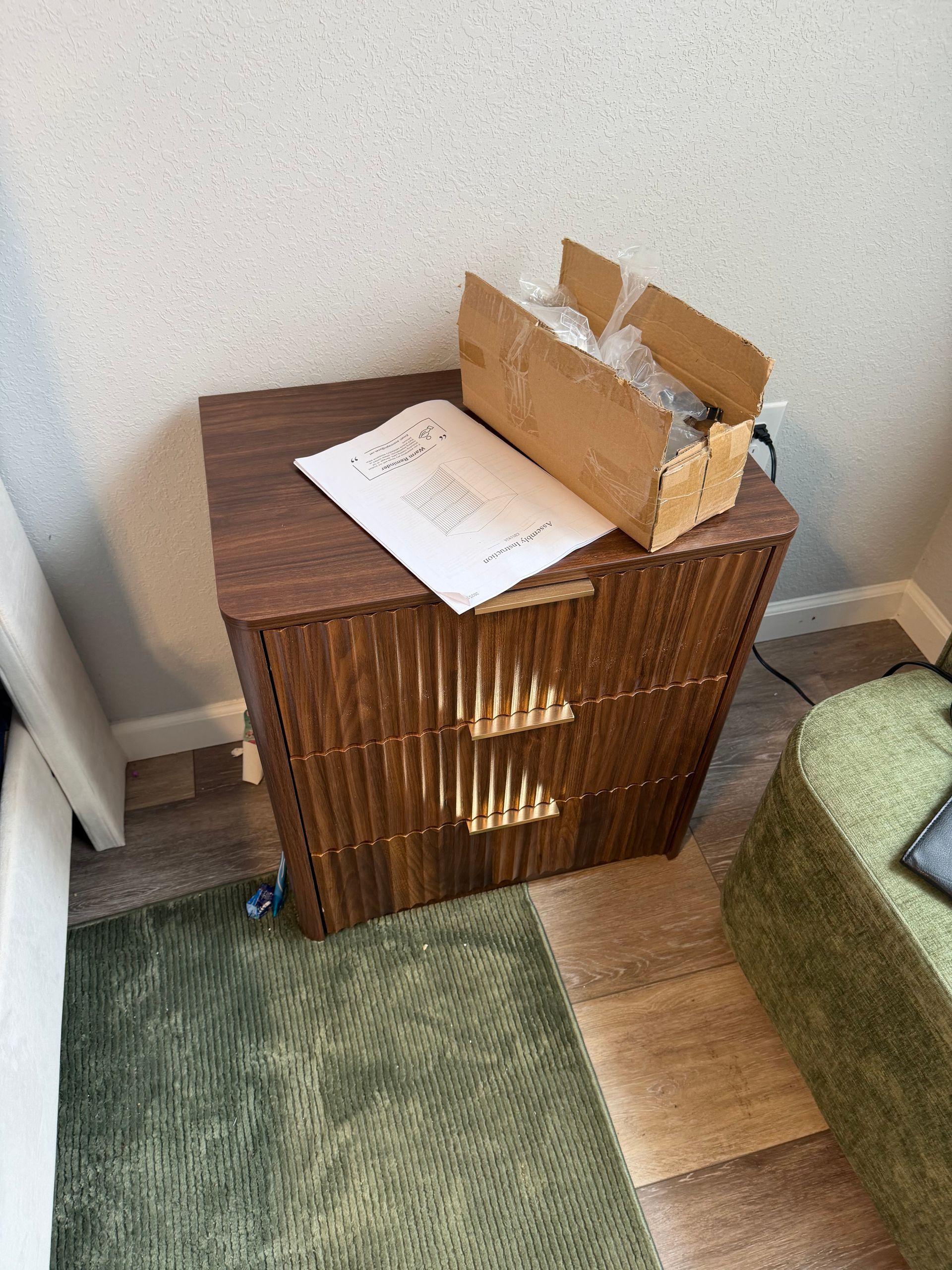 A wooden bedside table with a slatted front, a document, and a cardboard shipping box on top in a room with a green rug.