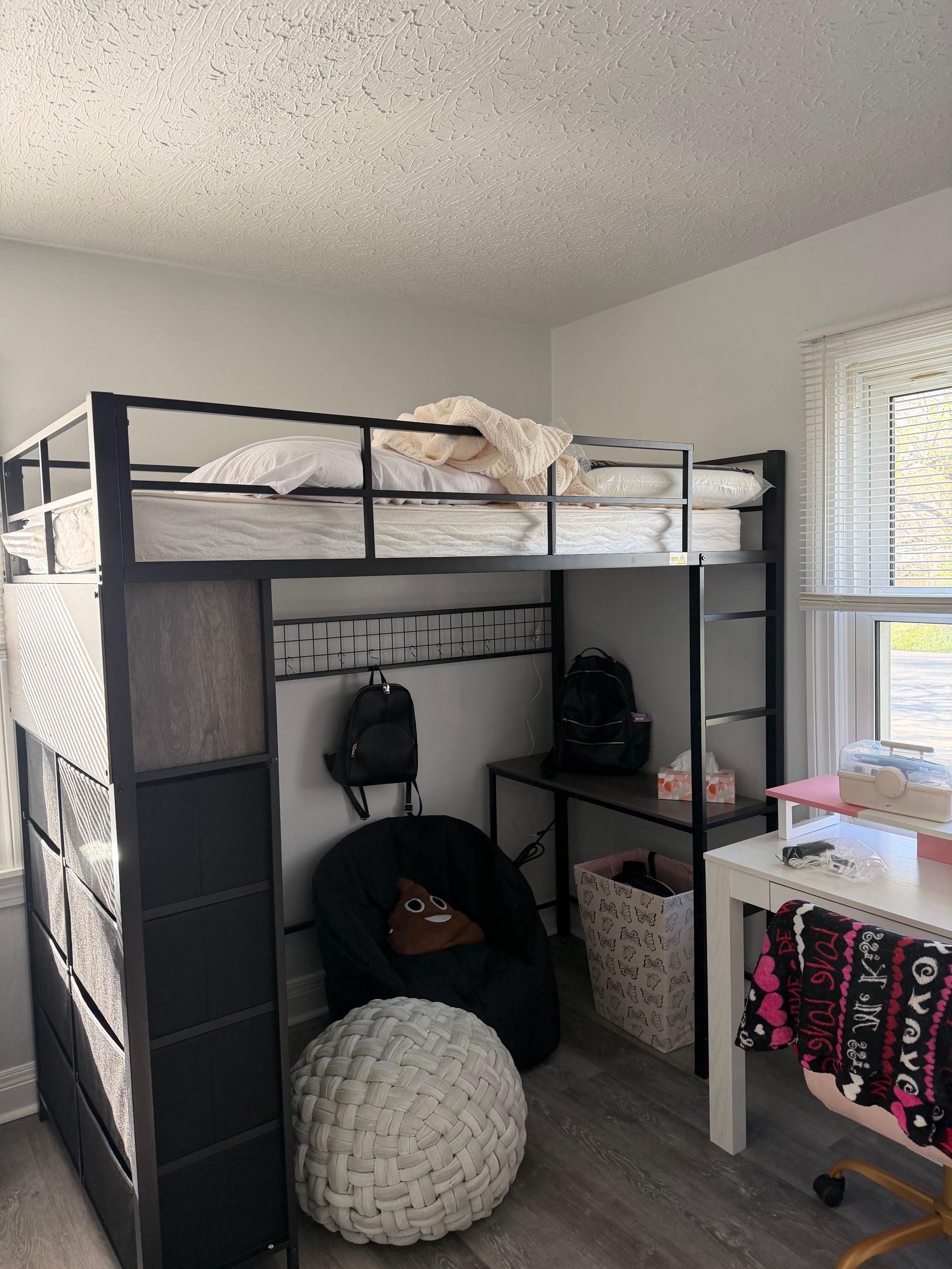 A black metal loft bed with a bed on top, a beanbag chair, a desk, and storage cubbies underneath in a bedroom.