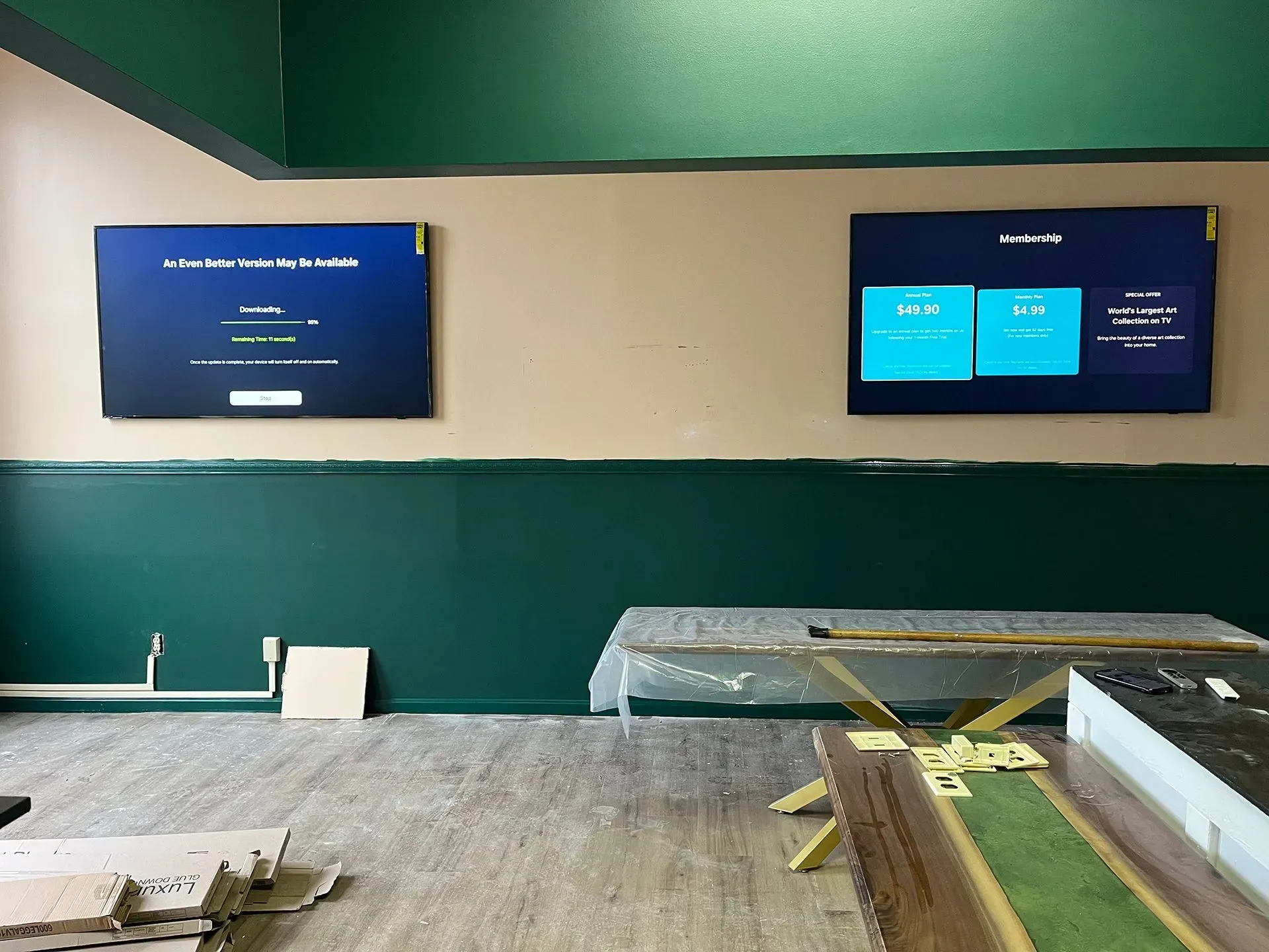 Two flat-screen monitors mounted on a beige and green wall in an unfinished room with construction materials present.