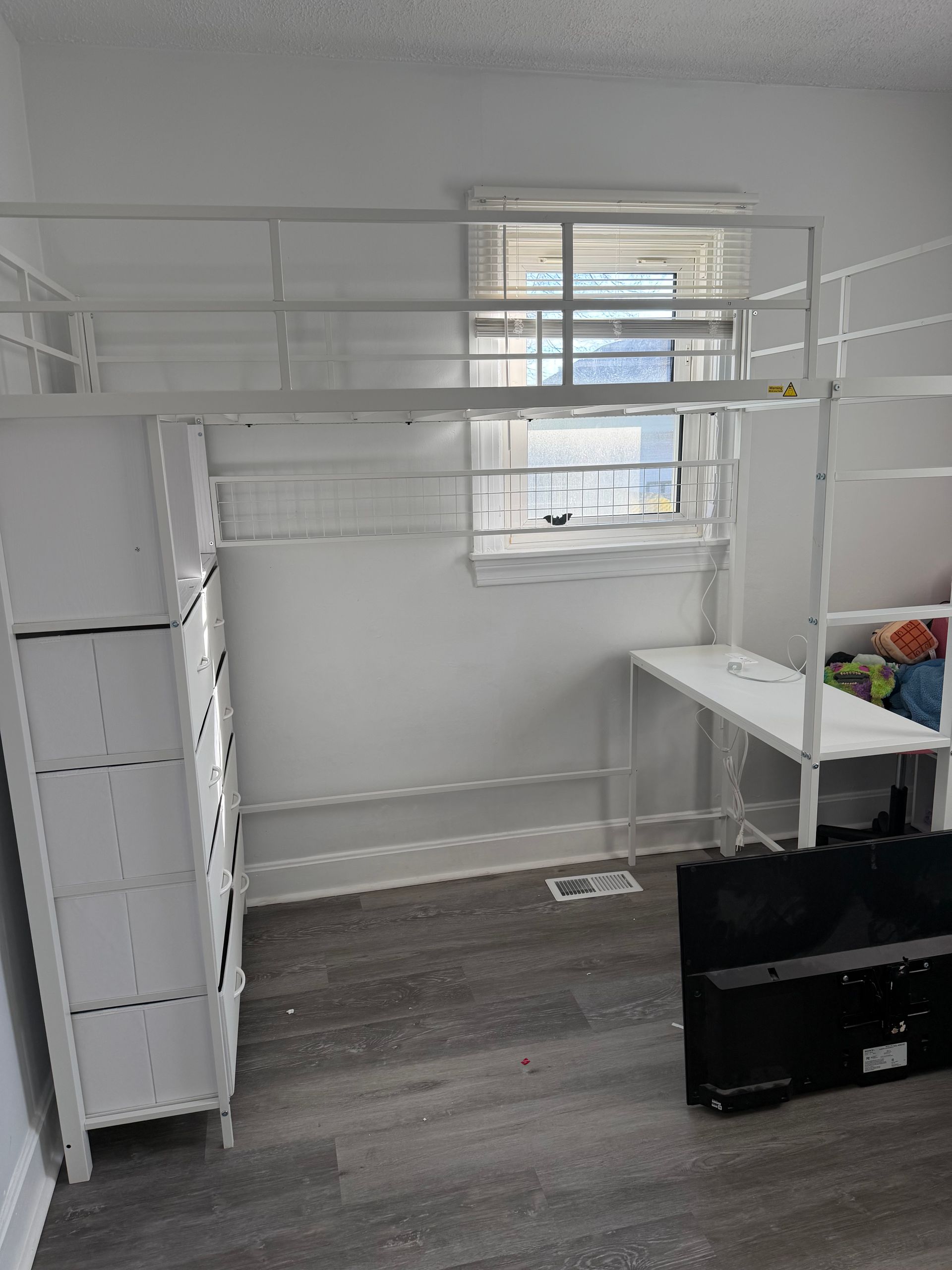 A white metal loft bed with an attached desk and a built-in dresser against a wall in a room with gray wood-style flooring.