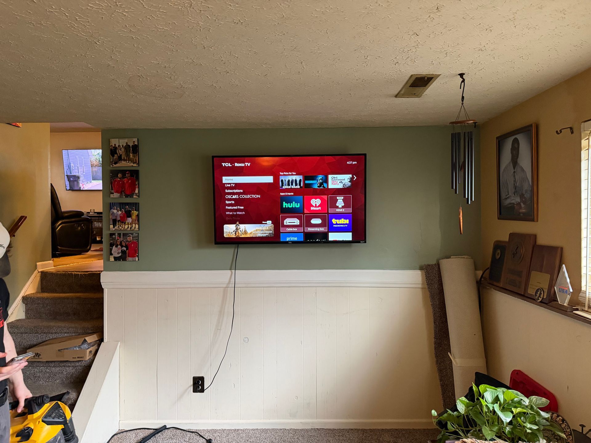 A wall-mounted TV displays a menu above wainscoting in a green room with stairs to the left and a framed photo on the right.