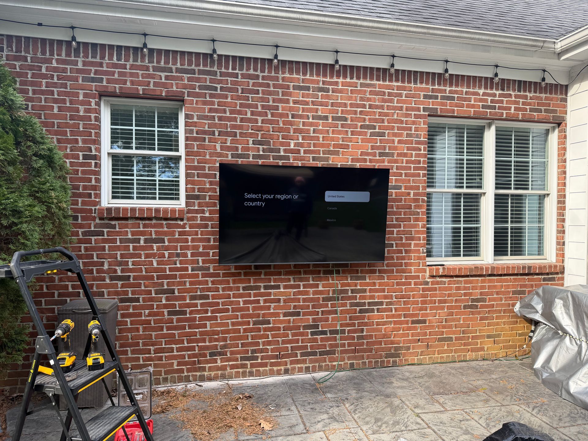 A television mounted on an outdoor brick wall between two windows, with a stepladder nearby on a patio.