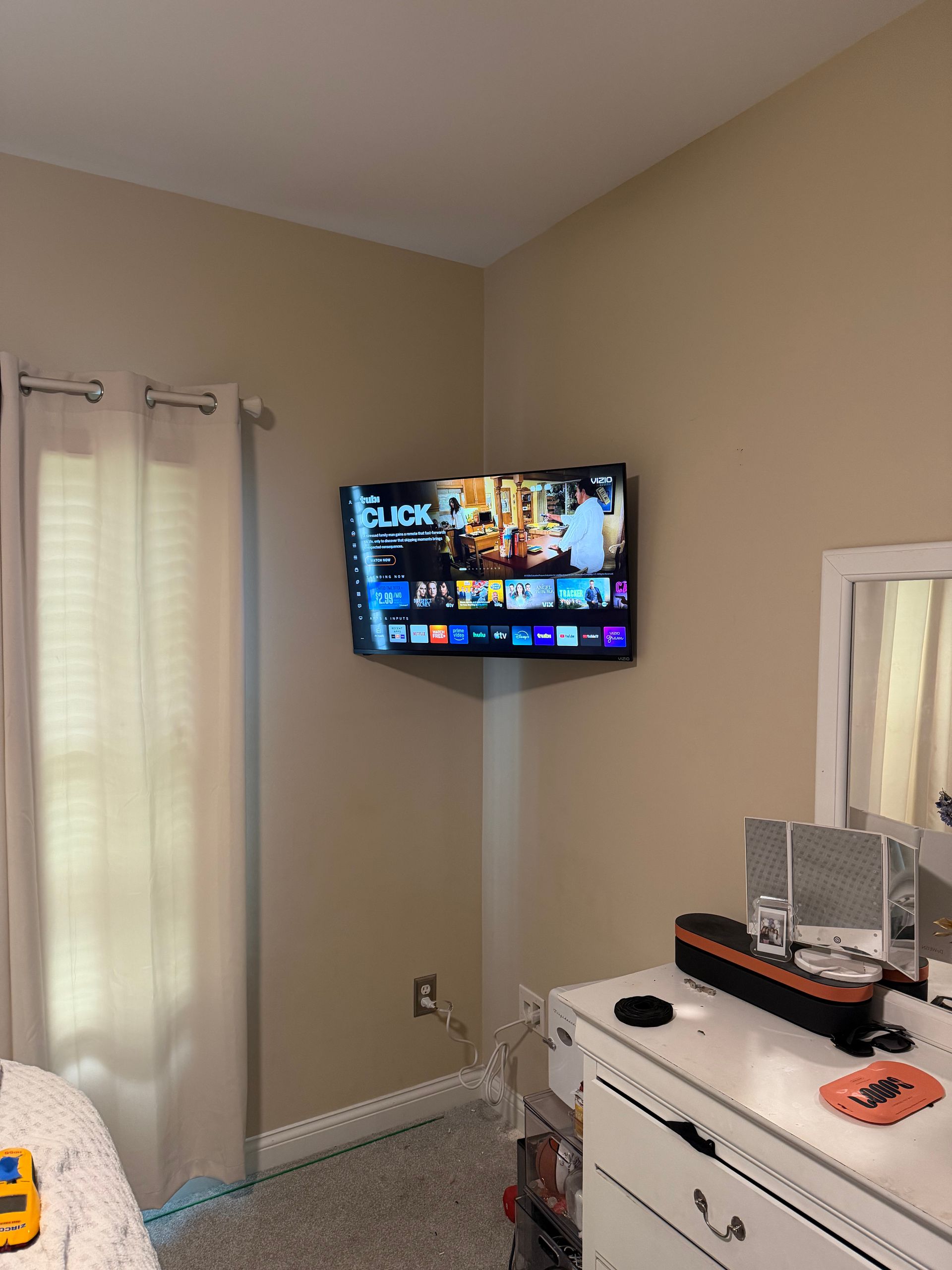 A wall-mounted TV in the corner of a beige room, with a dresser and mirror below it next to a window with curtains.