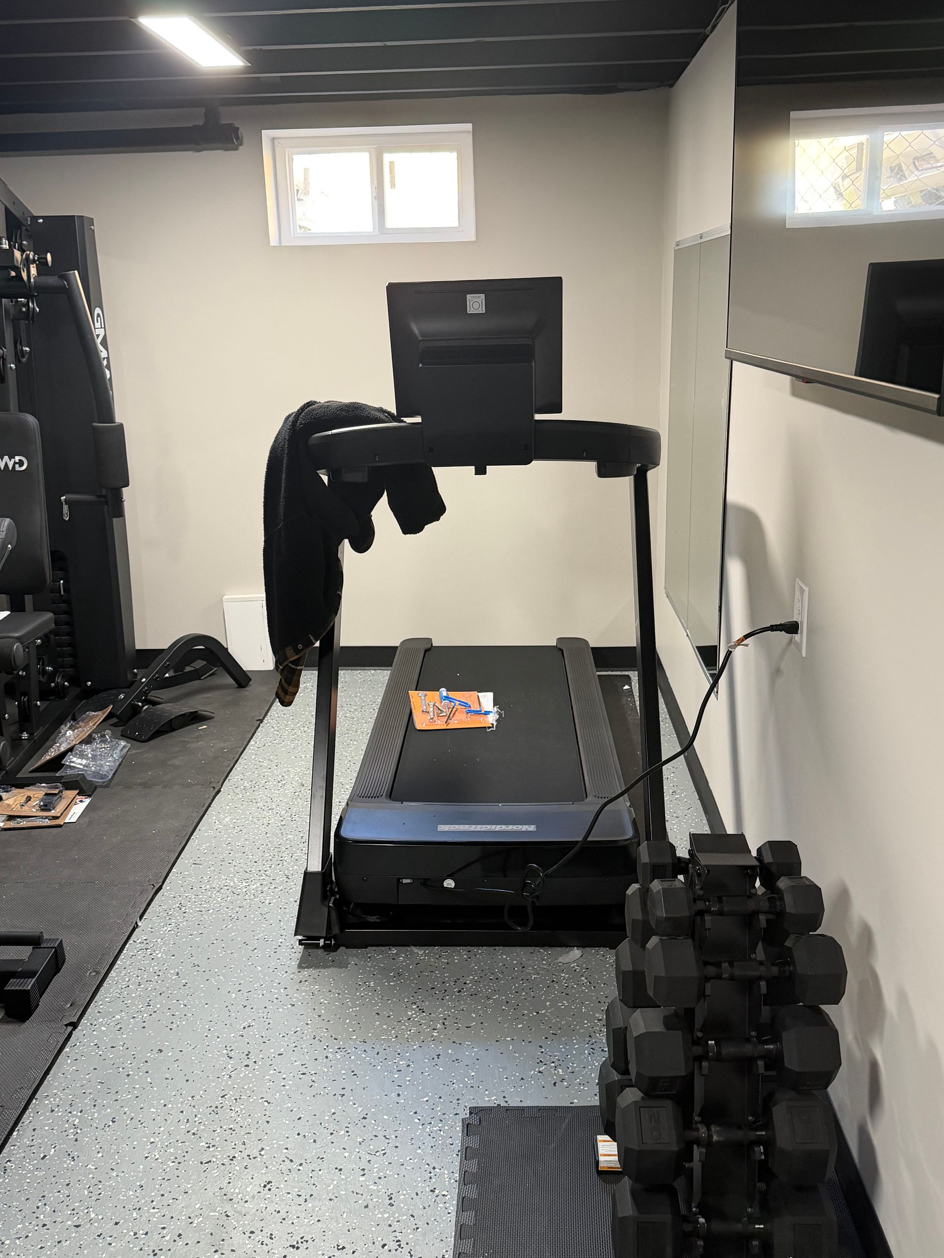 A home gym featuring a treadmill, a dumbbell rack, and a weight station on a grey speckled floor against light walls.