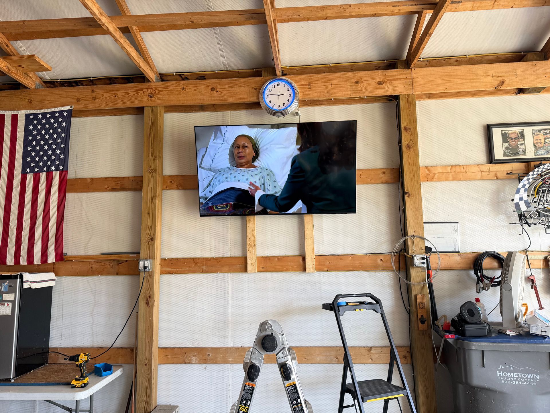 A television mounted on wooden garage wall studs showing a person in a hospital bed, with a flag and tools nearby.
