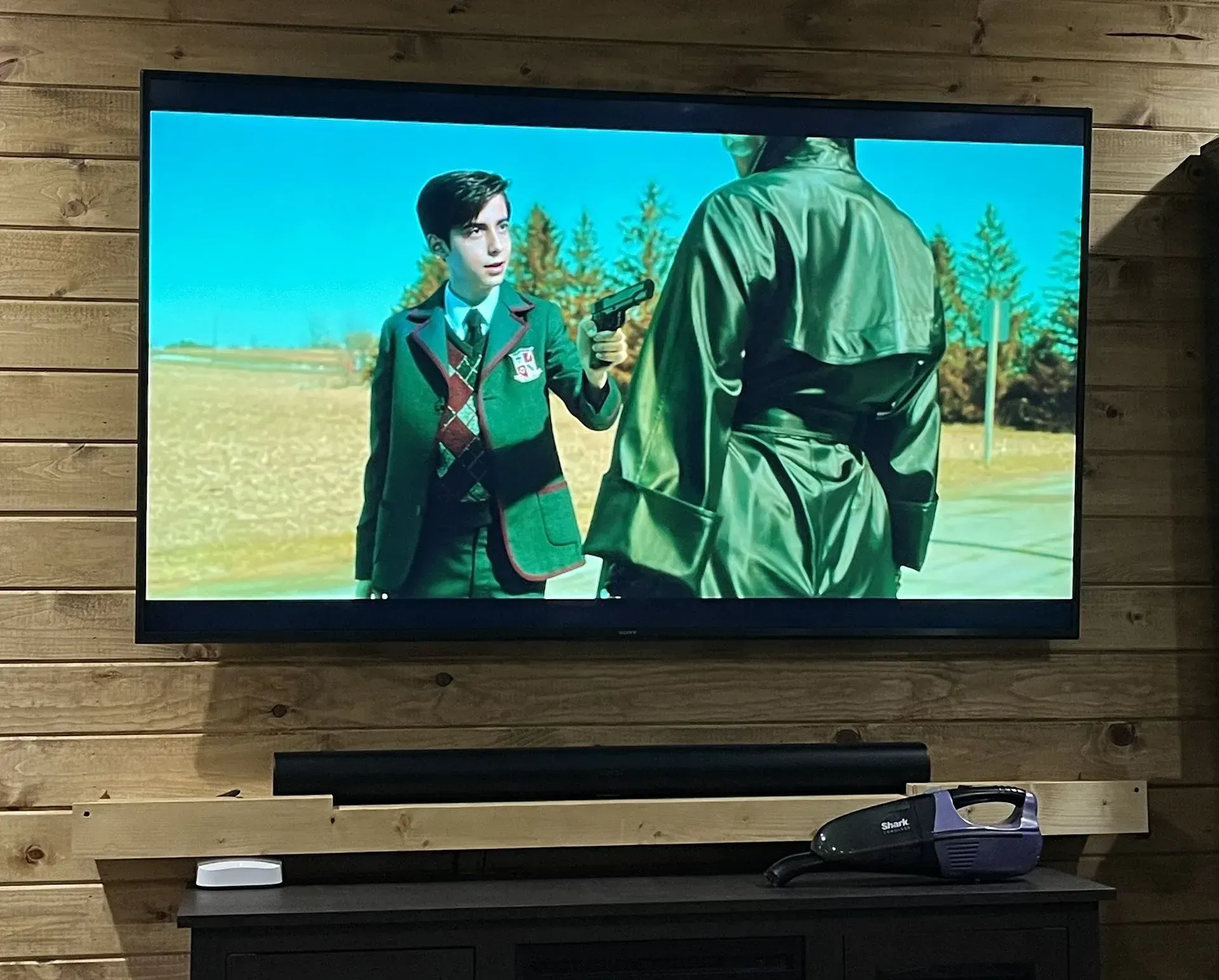 A TV screen mounted on a wood wall displays a scene with a person in a green blazer holding a gun toward another person.