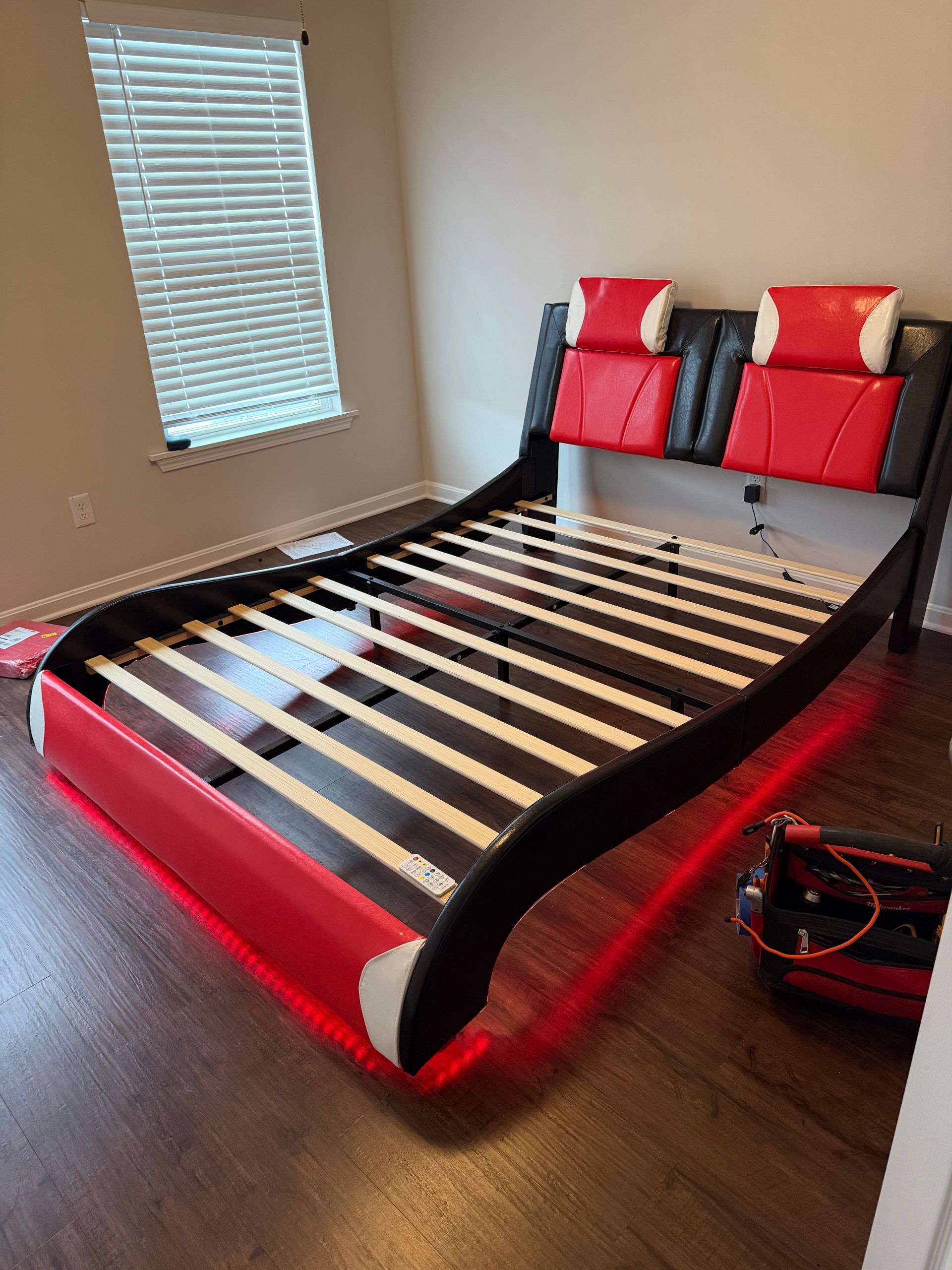 A modern red and black upholstered bed frame with headrest cushions and wooden slats in a room with a window.
