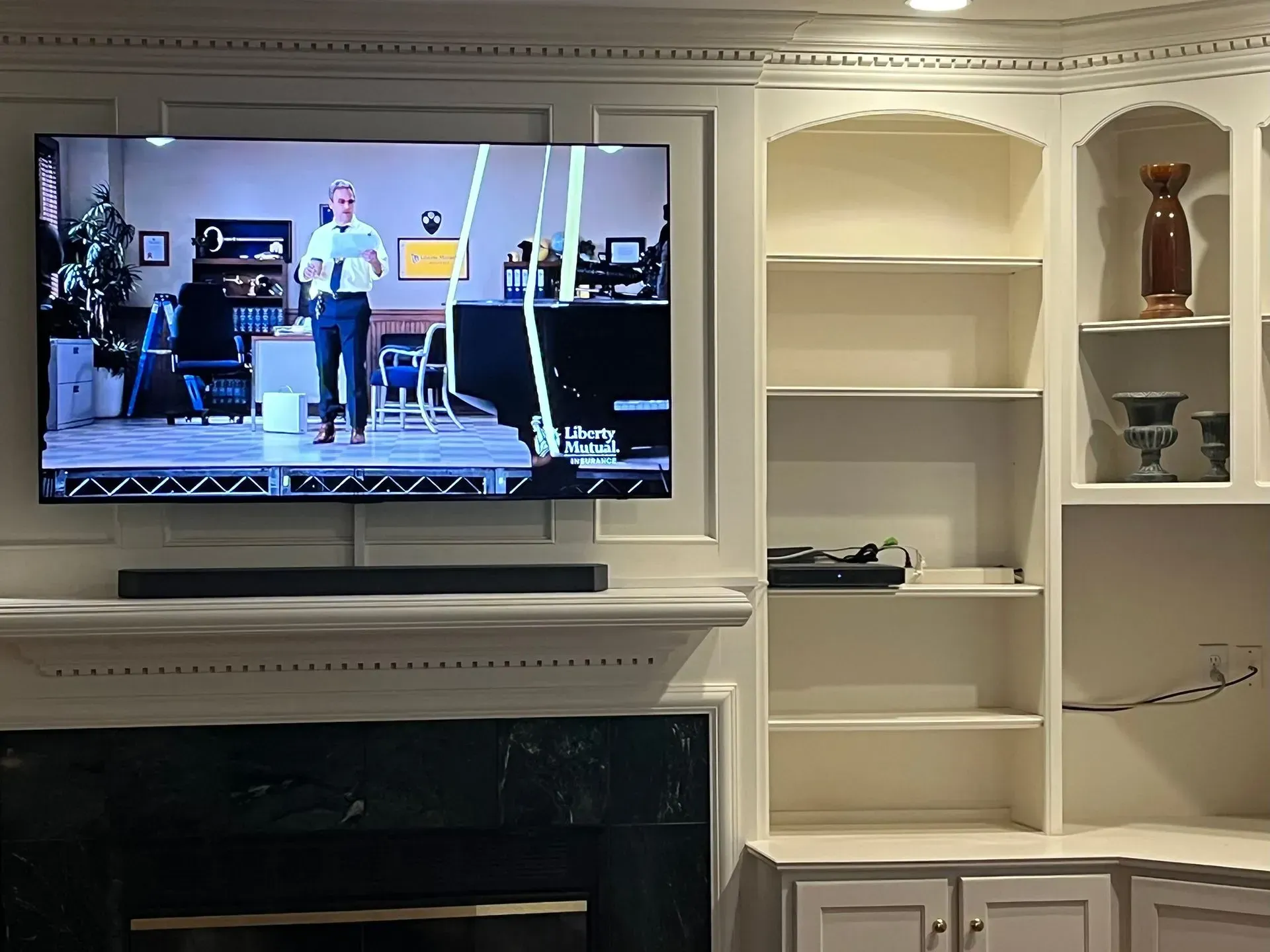 A wall-mounted TV displaying a person giving a presentation above a fireplace, next to white built-in bookshelves.
