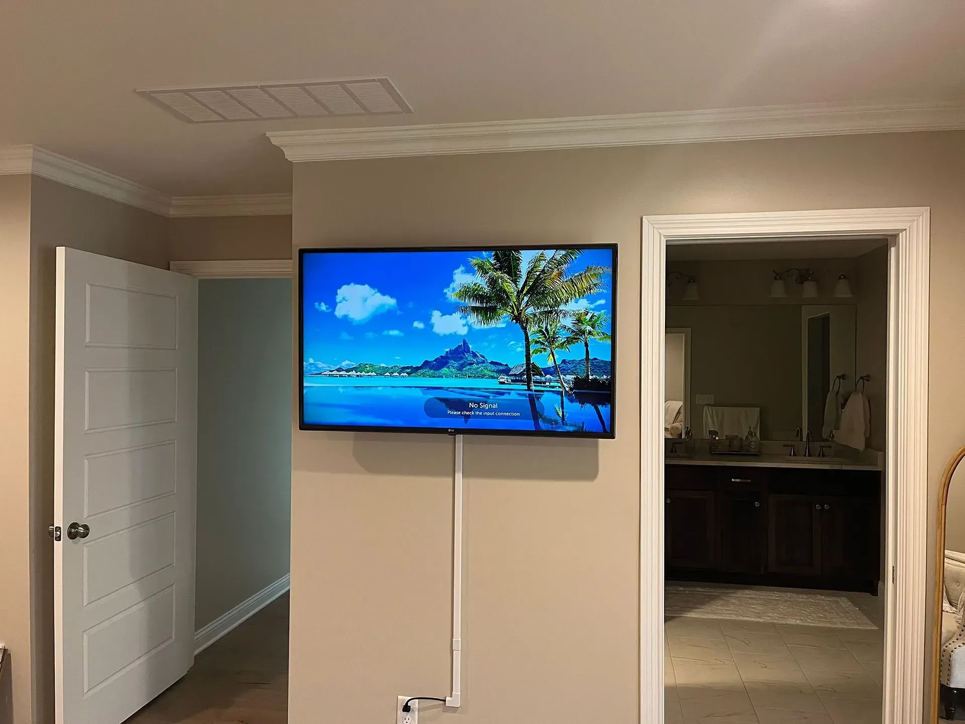 A flat-screen TV displaying a beach scene is mounted on a beige wall between two doorways, with white cord covers below.