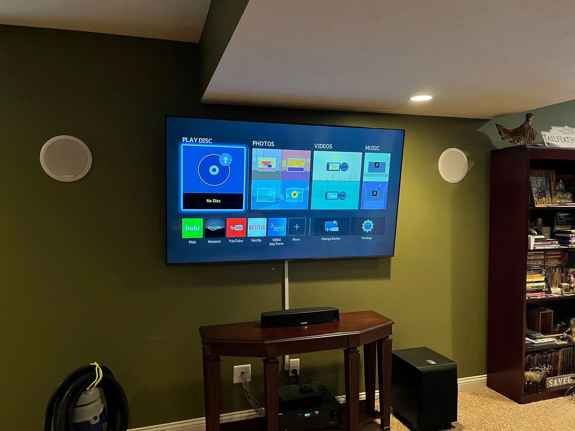 A flat-screen TV mounted on a green wall with two speakers, a console table below, and a shelving unit to the right.