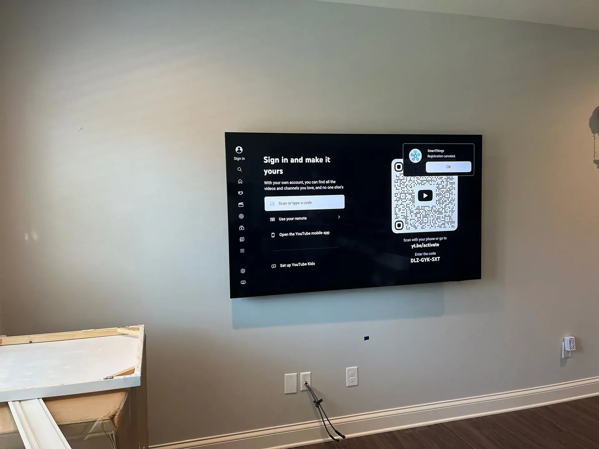 A flat-screen TV mounted on a light gray wall displaying a YouTube settings menu with visible cables below.