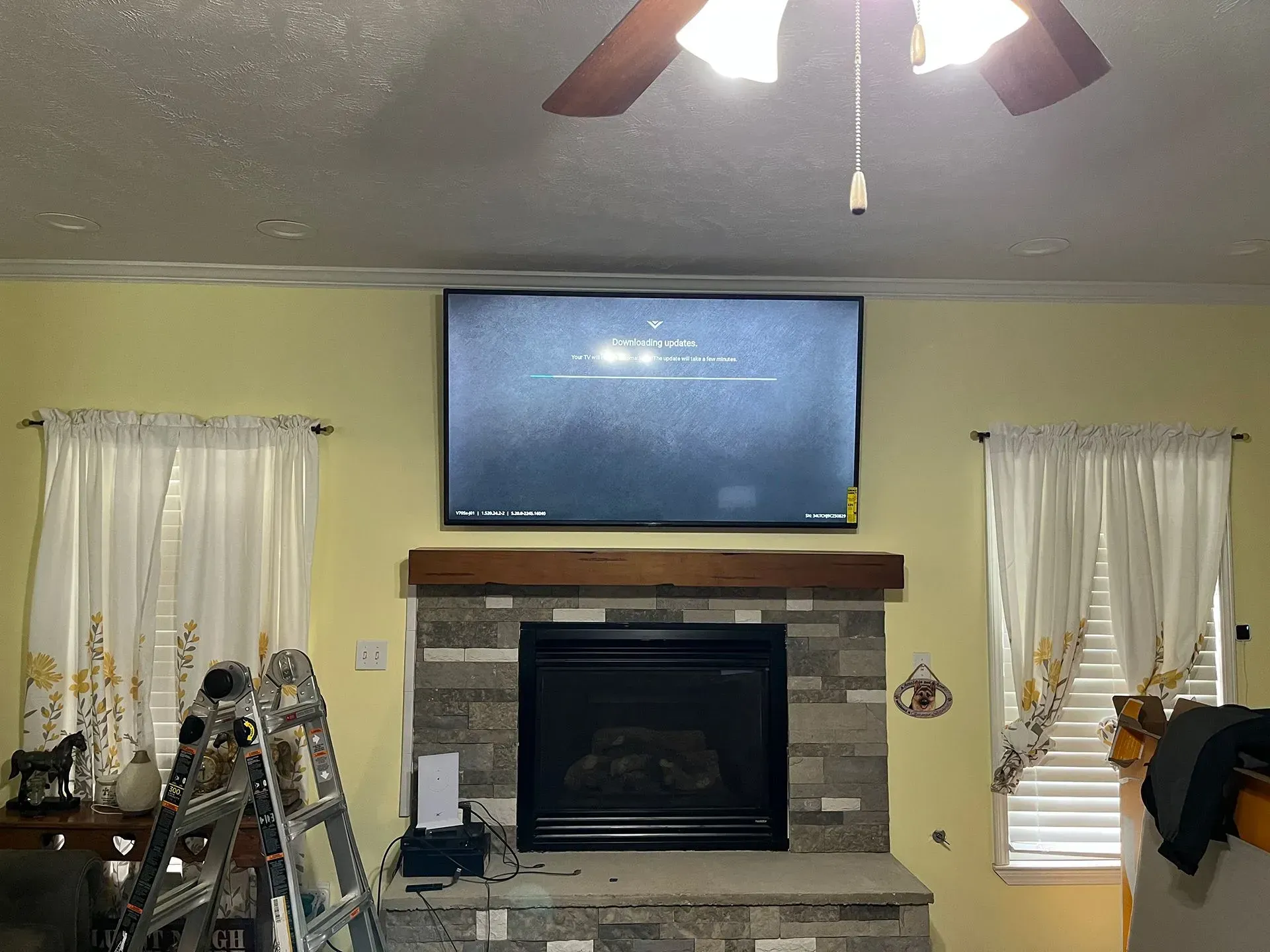 A large television mounted above a stone fireplace in a room with light yellow walls and two windows with white curtains.