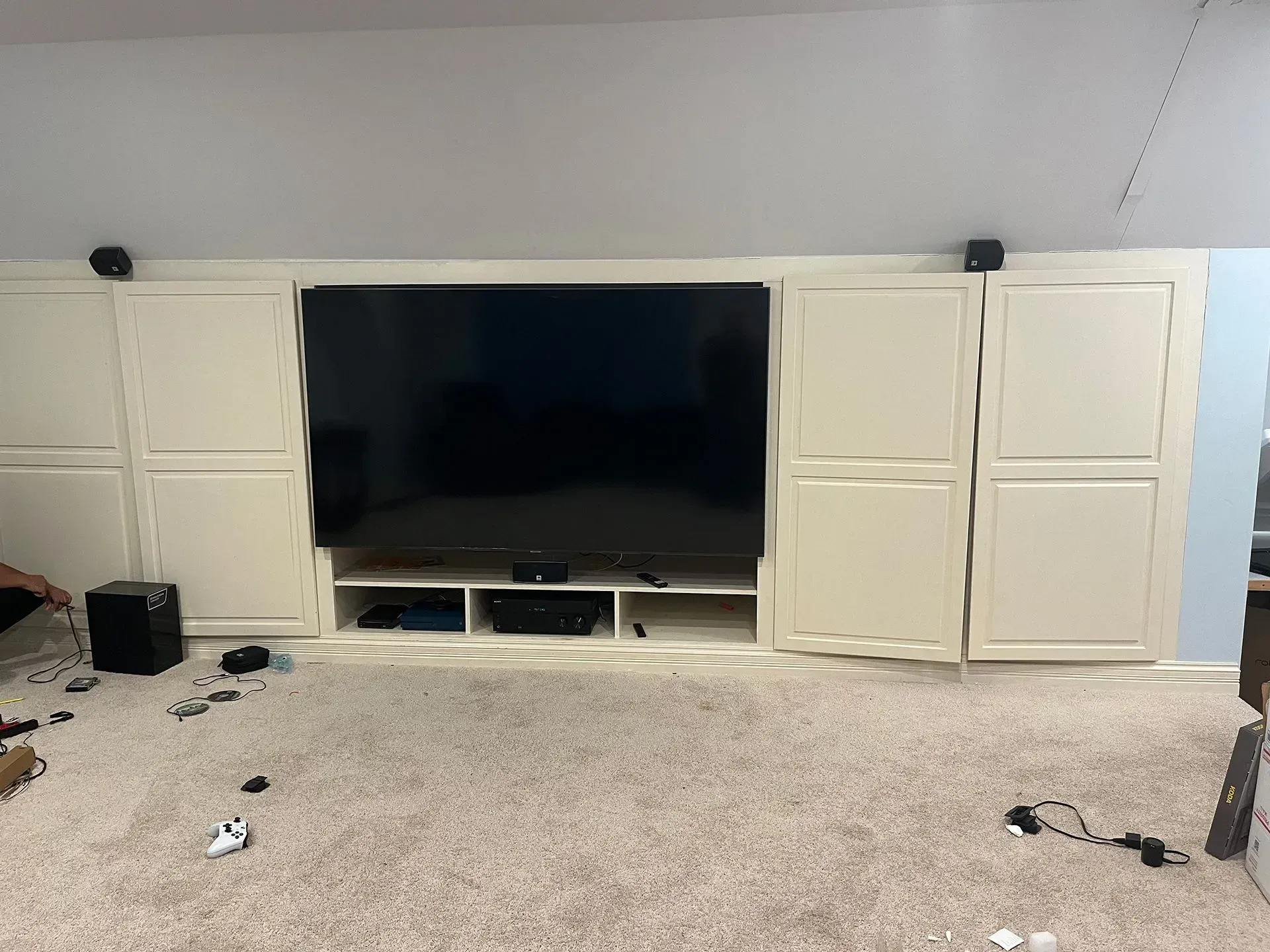 A wall-mounted TV flanked by white cabinet doors with open shelving below, set against a room with beige carpet.