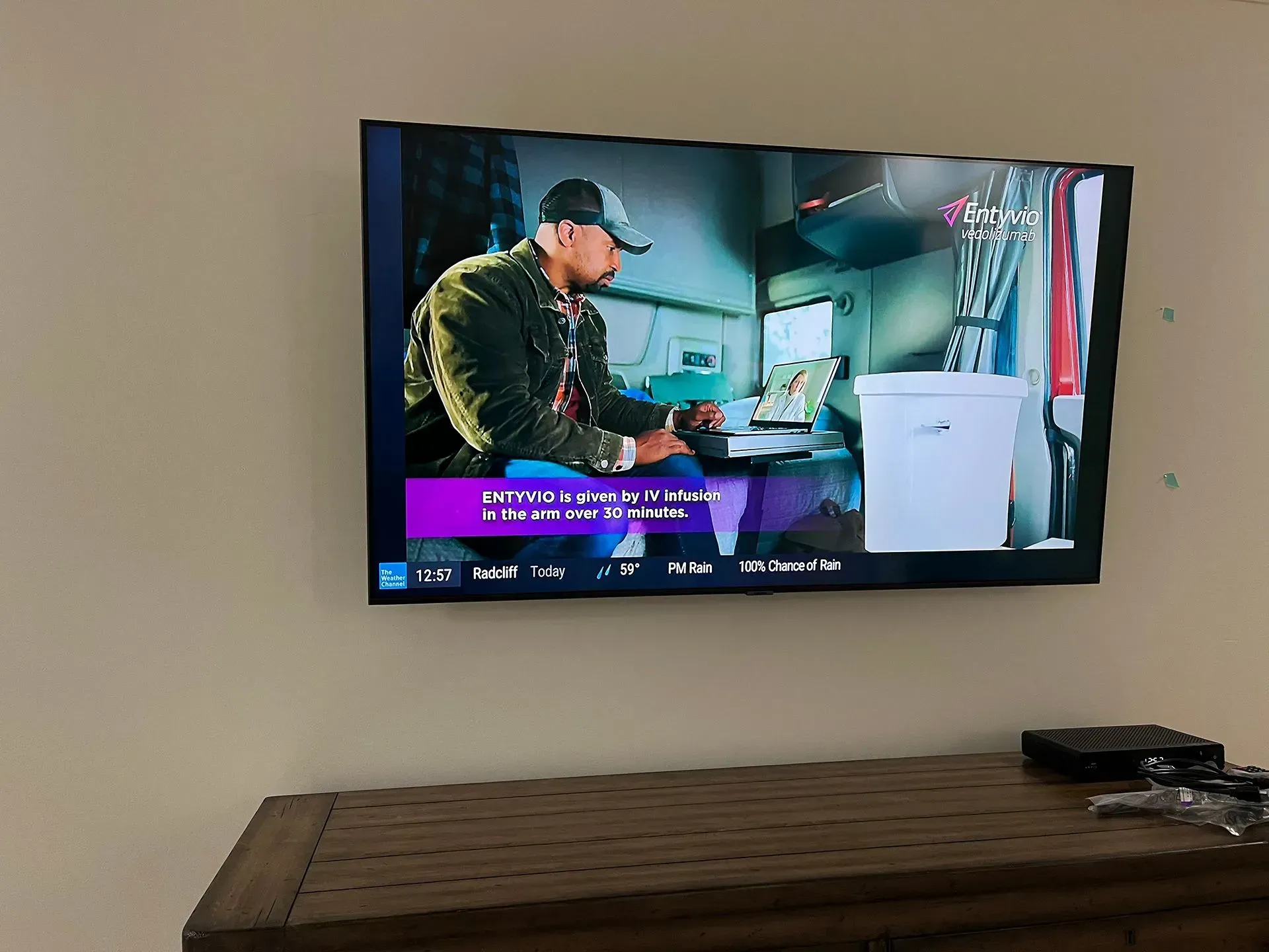 A wall-mounted television displays a person wearing a cap, seated at a desk near a white container in a room.
