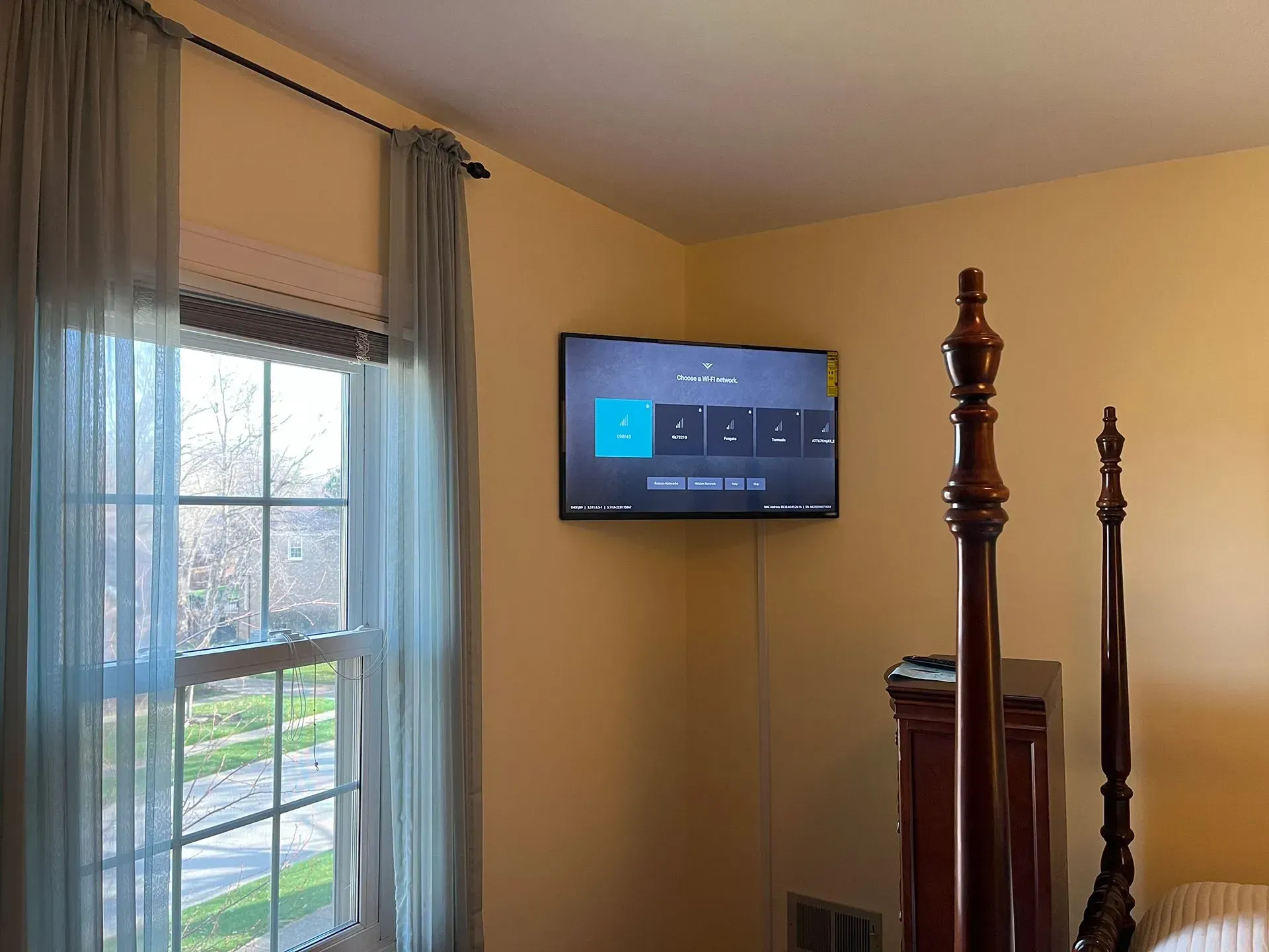 A flat-screen TV mounted on a tan wall in a bedroom next to a window with light blue curtains.