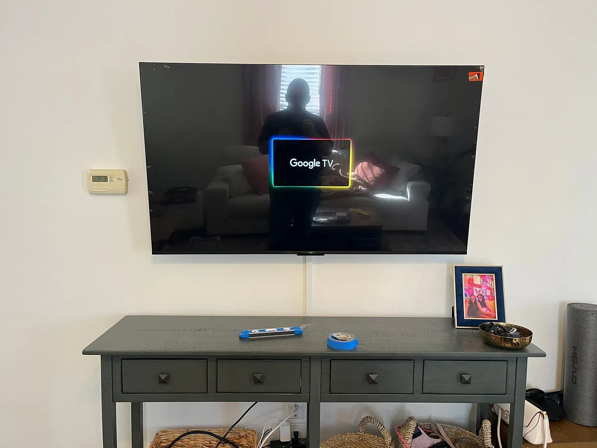 A TV mounted on a wall displaying the Google TV logo above a gray wooden console table with drawers.