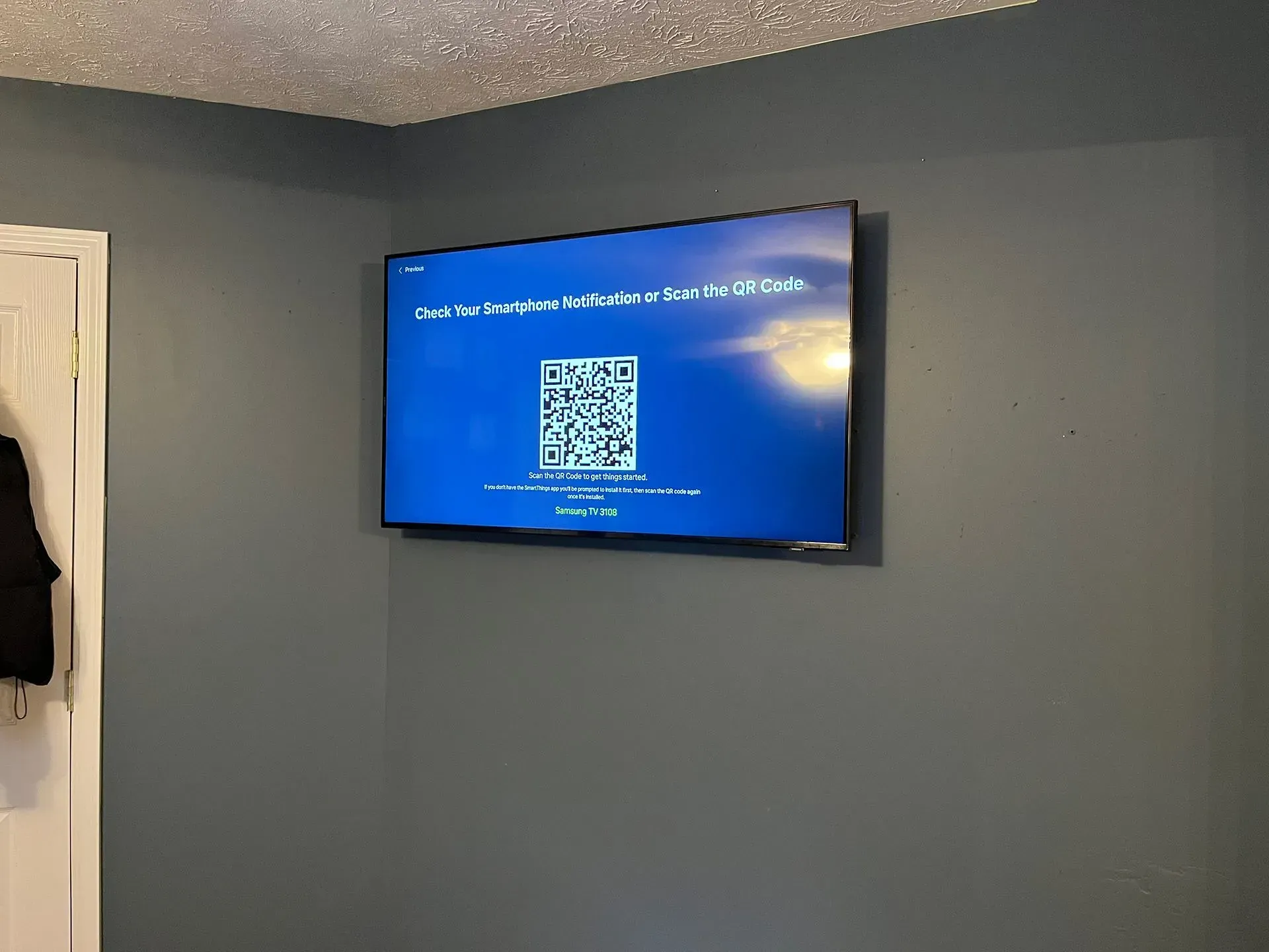 A flat-screen TV mounted on a dark gray wall displaying a QR code for device setup on a blue screen.
