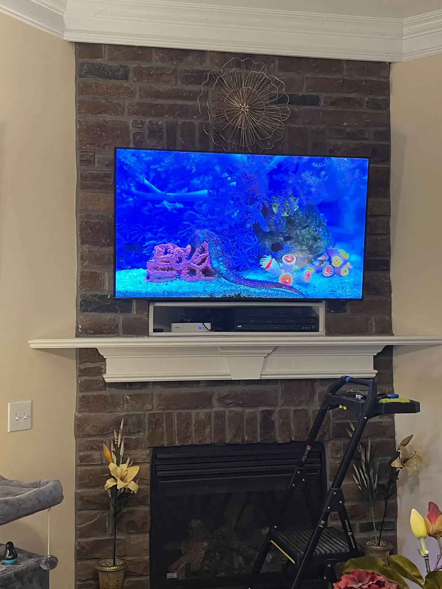 A television mounted above a fireplace on a brown brick wall, with a black stepladder placed in front.