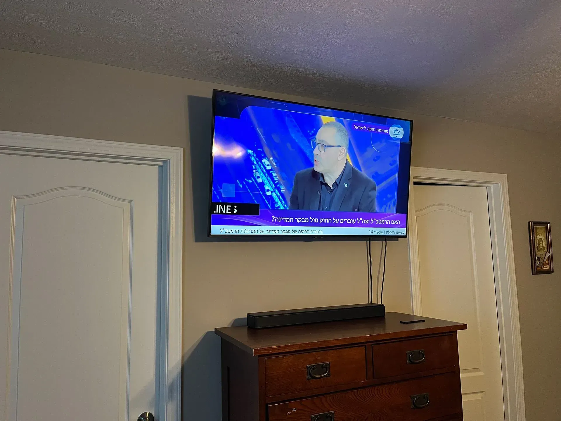 A flat-screen TV mounted on a wall above a wooden dresser, displaying a news broadcast between two white doors.