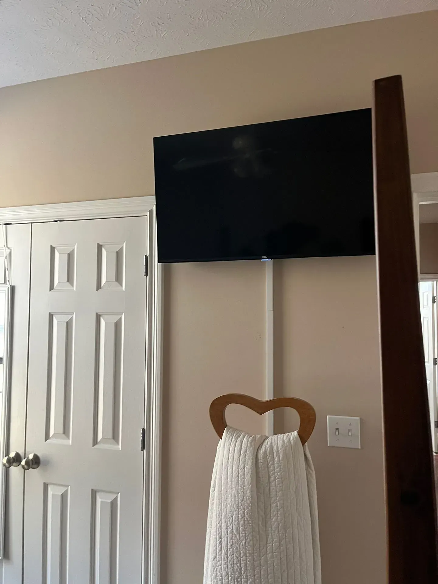 A flat-screen TV mounted on a wall above a white towel hanging from a heart-shaped wooden hook.