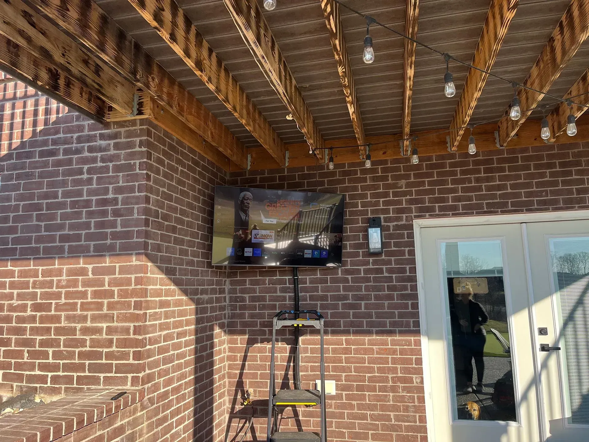 A flat-screen television mounted on a red brick exterior wall under a wooden patio deck with string lights.