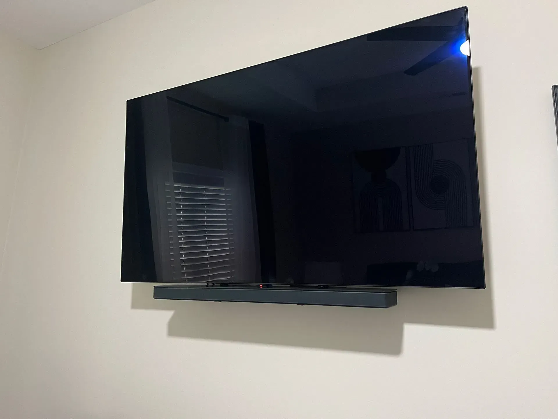 A large, dark-screen television is mounted on a plain light wall with a slim black soundbar positioned directly below it.