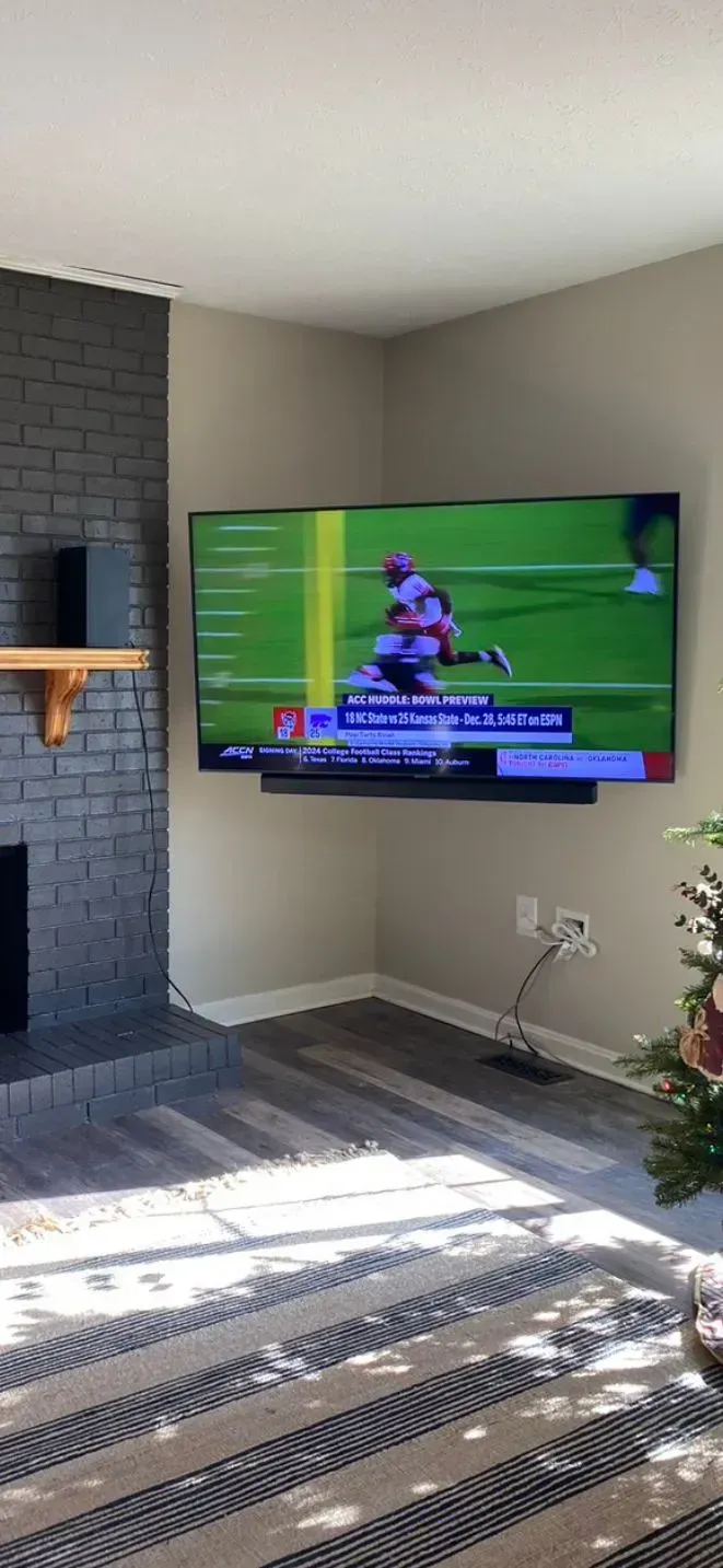 A large flat-screen TV mounted on a beige wall next to a dark painted brick fireplace, displaying a football game.