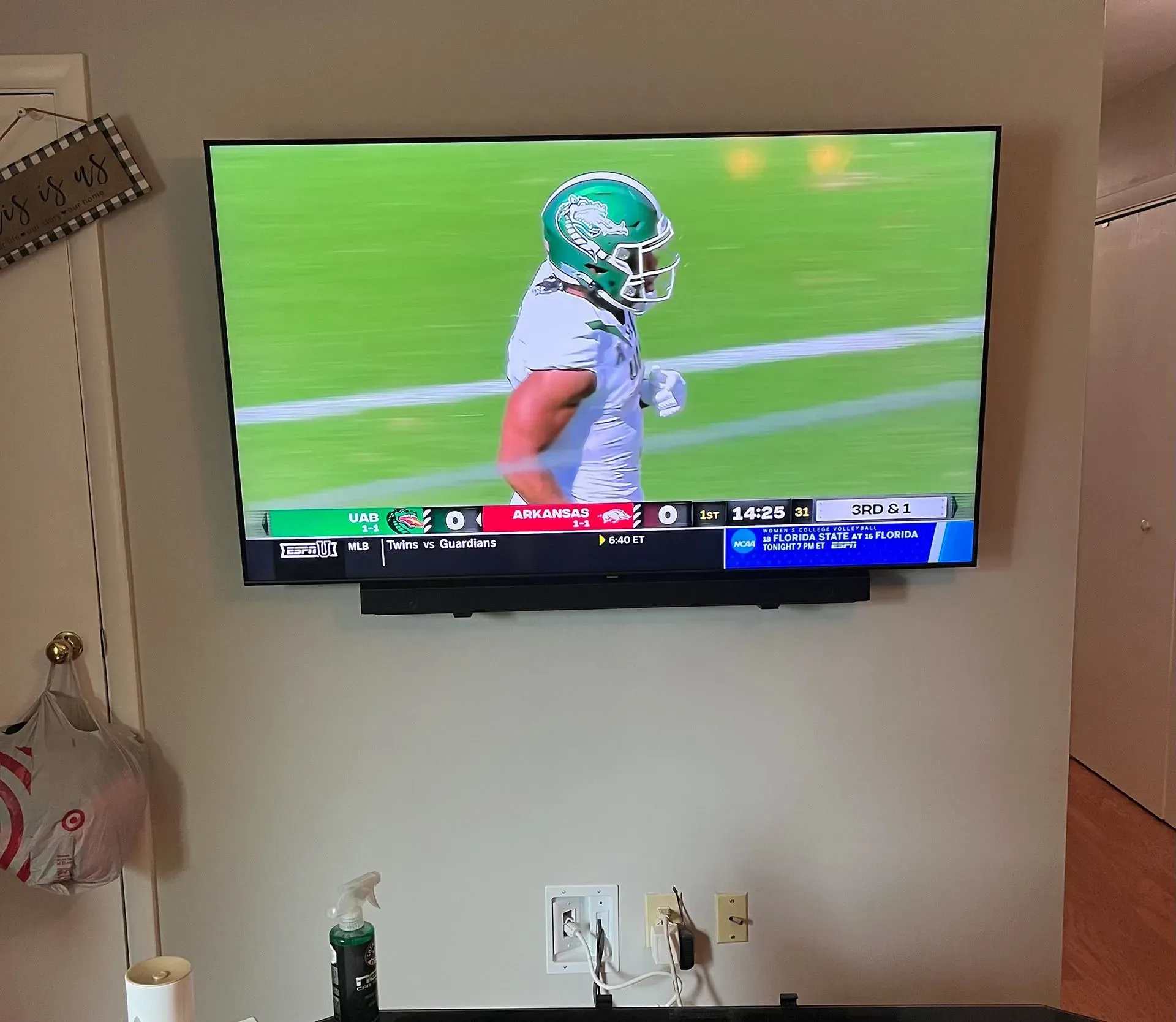 A wall-mounted television displays a football player in a white uniform with a green helmet on a field during a game.