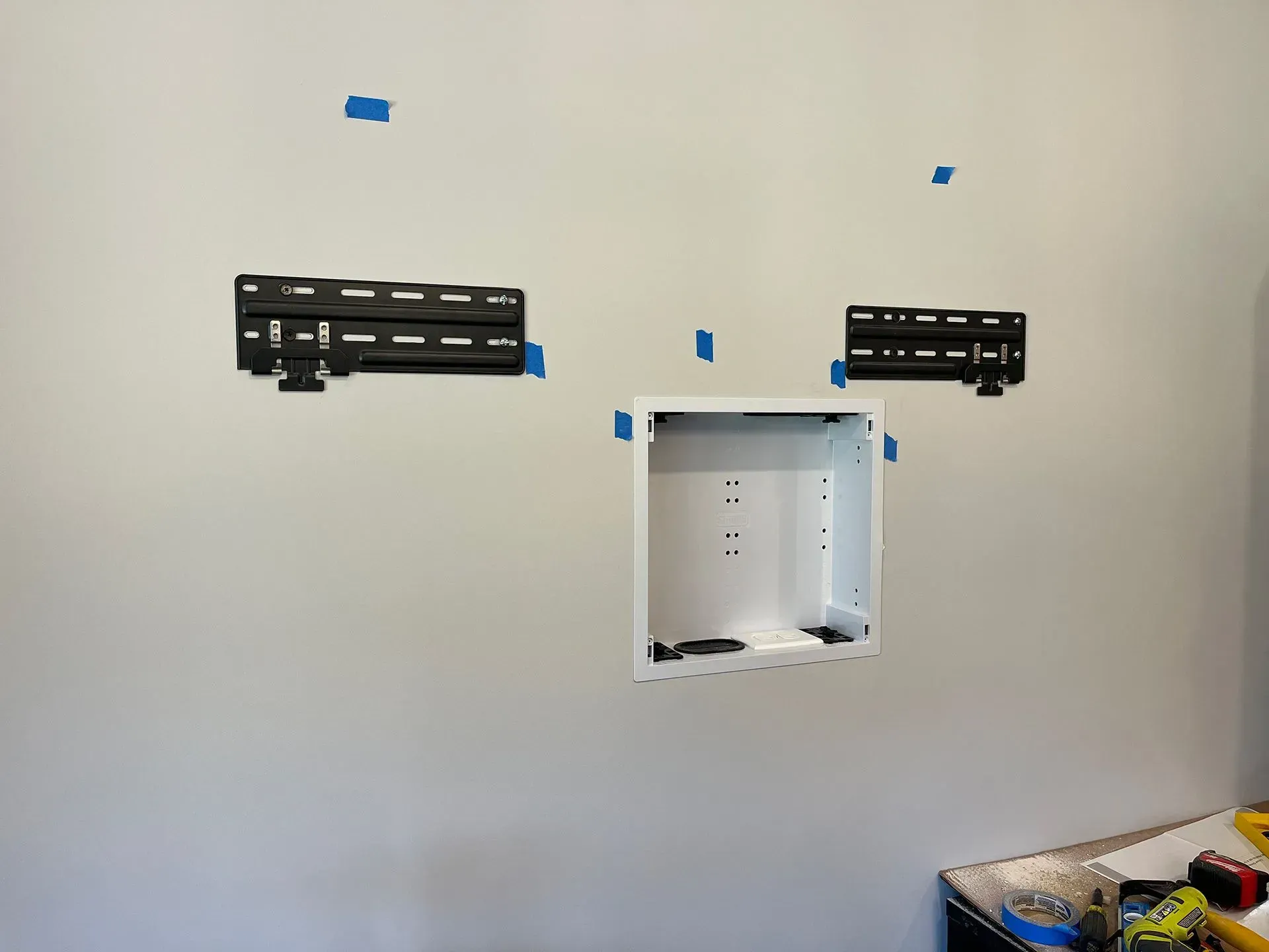 Two TV mounting brackets and a recessed media box installed on a light-colored wall with blue painter's tape markers.