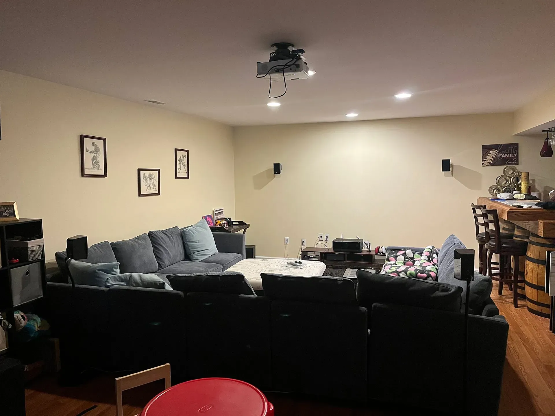 A basement living room features a dark sectional sofa, a projector mounted on the ceiling, and a home bar area.