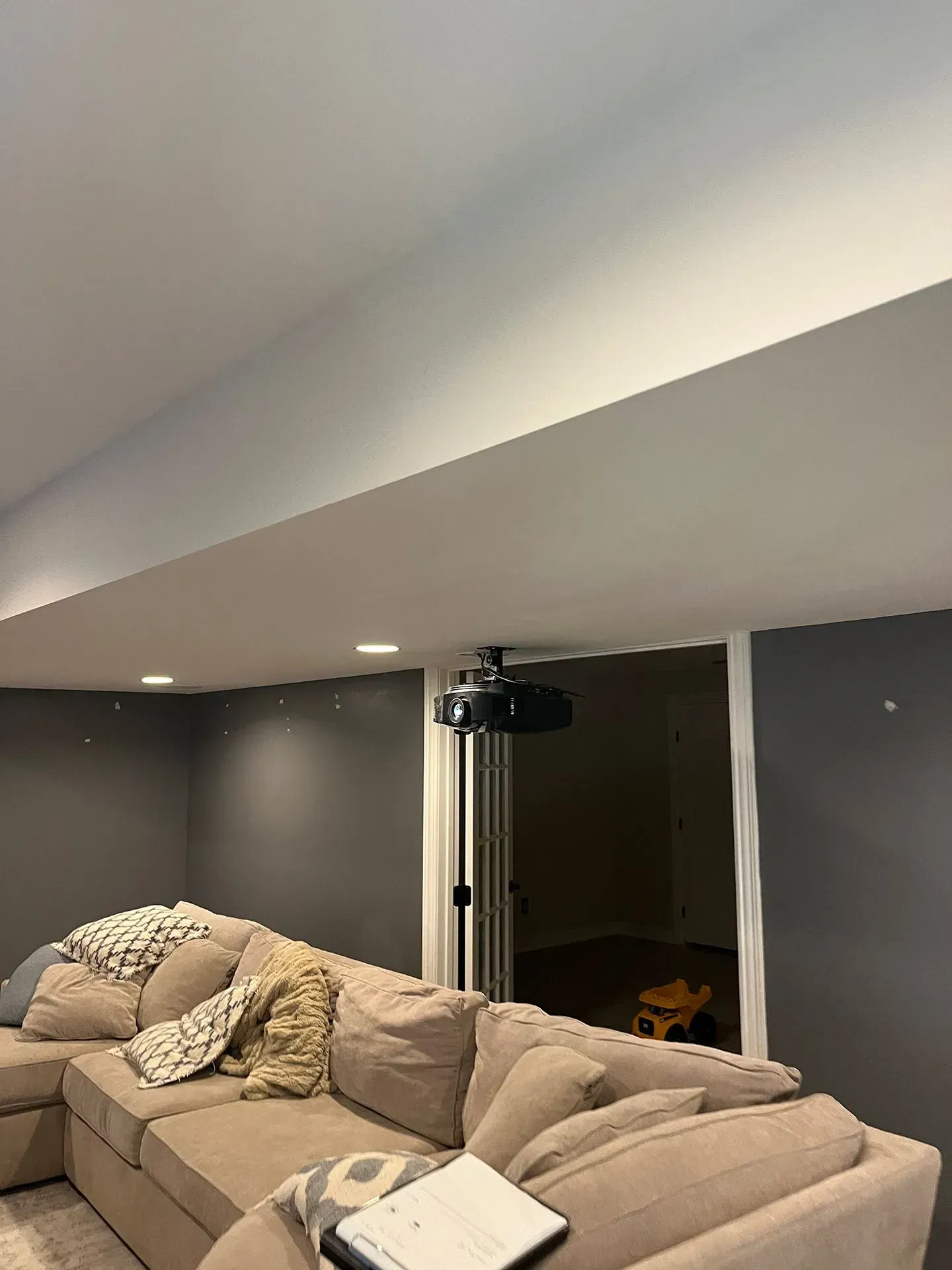 A projector mounted on a tall stand in front of a beige sofa in a room with gray walls and recessed ceiling lighting.