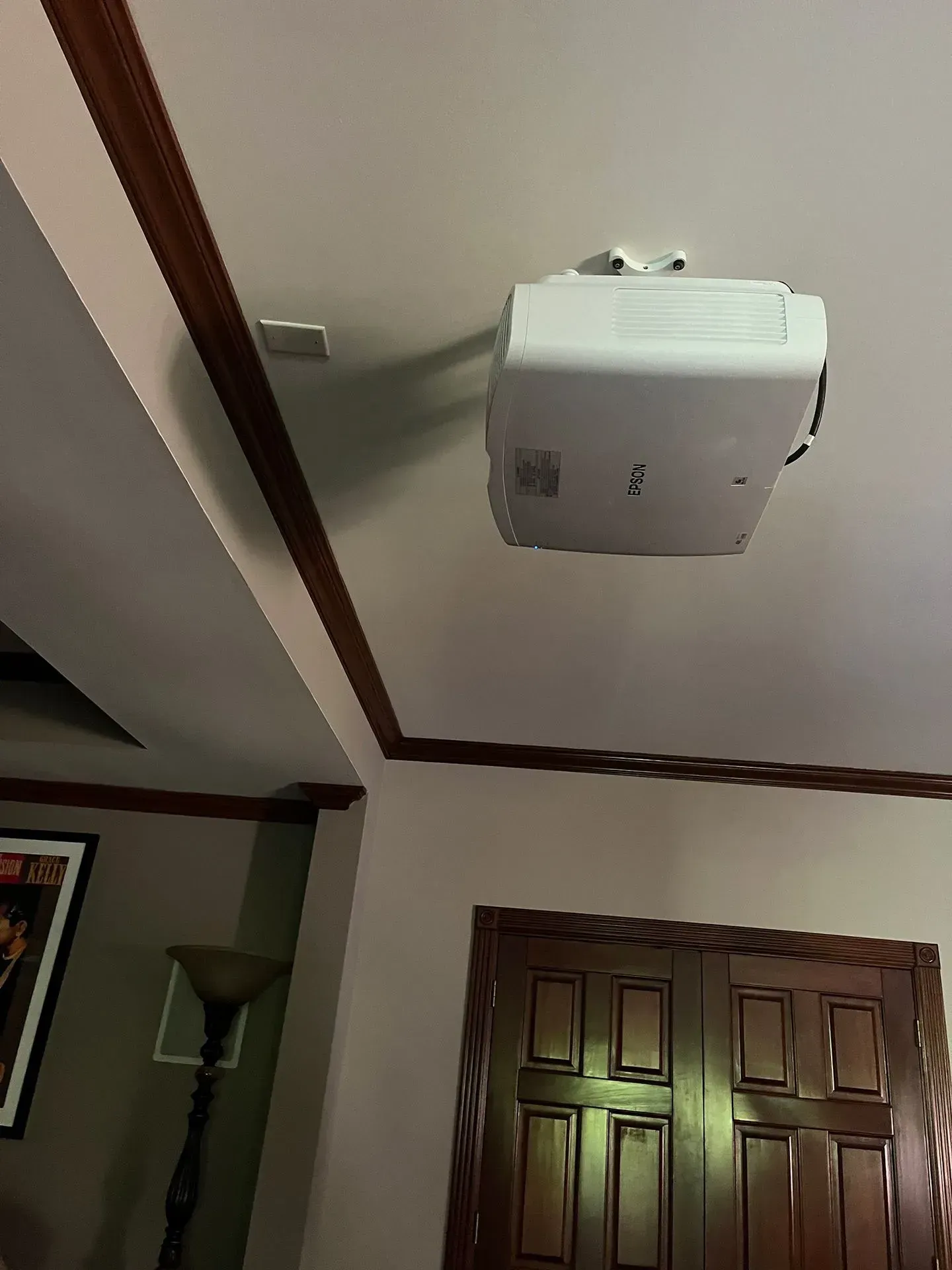 A white digital projector is mounted to a ceiling in a room with dark wood trim and a wooden door.