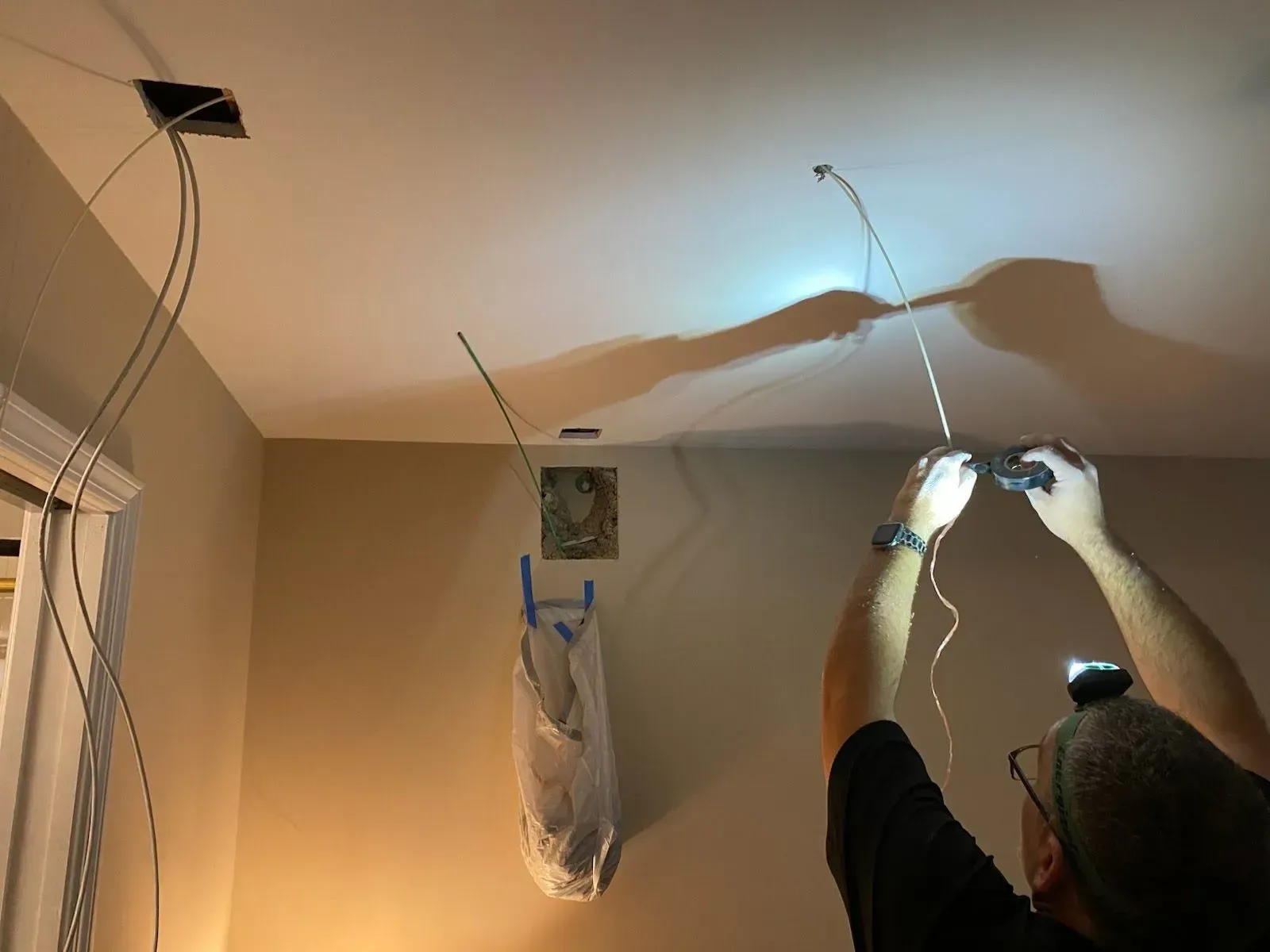 A person works on electrical wiring hanging from holes in a room ceiling while wearing a headlamp.