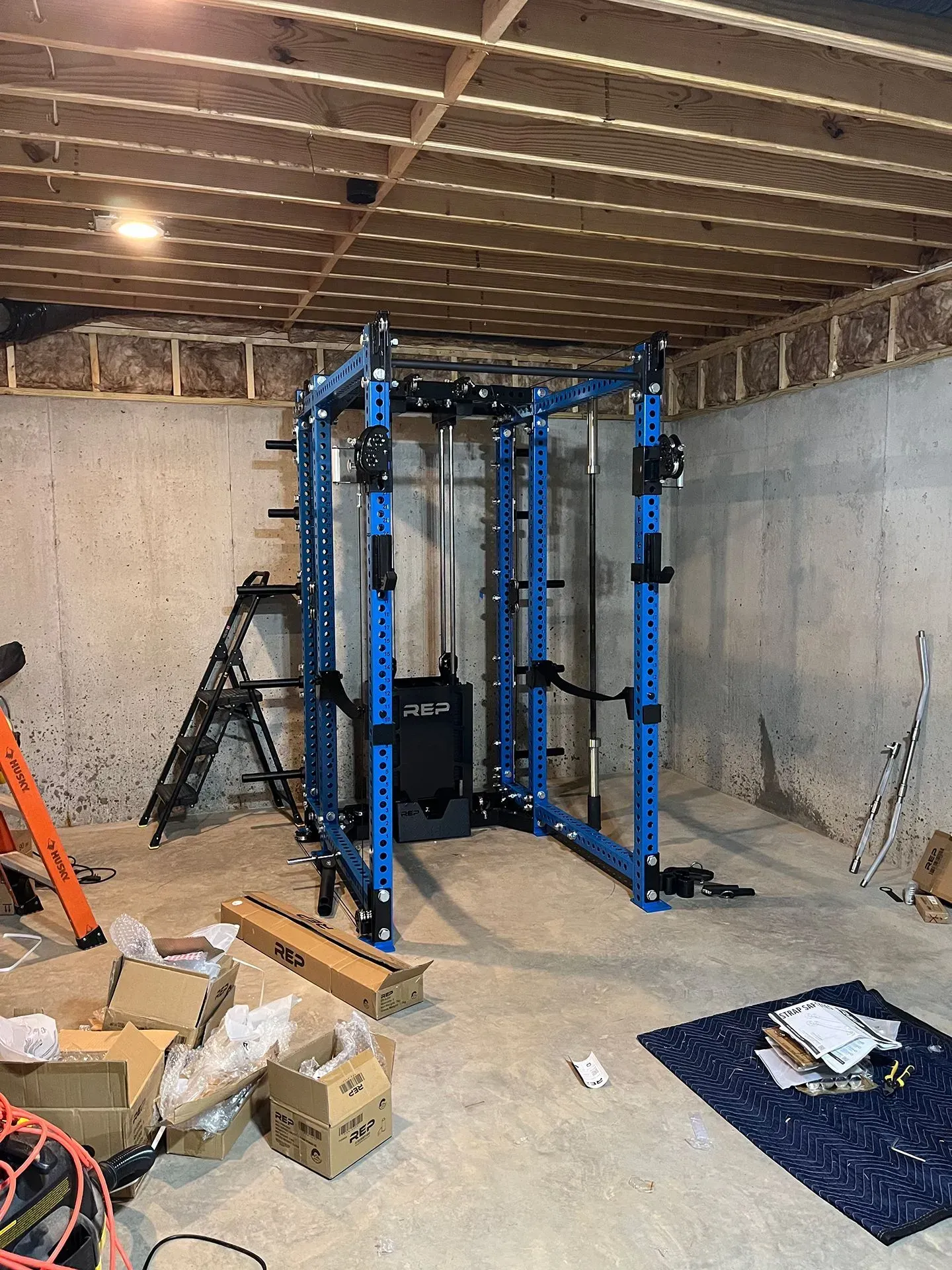 A blue gym power rack stands in an unfinished concrete basement, with scattered cardboard boxes on the floor.