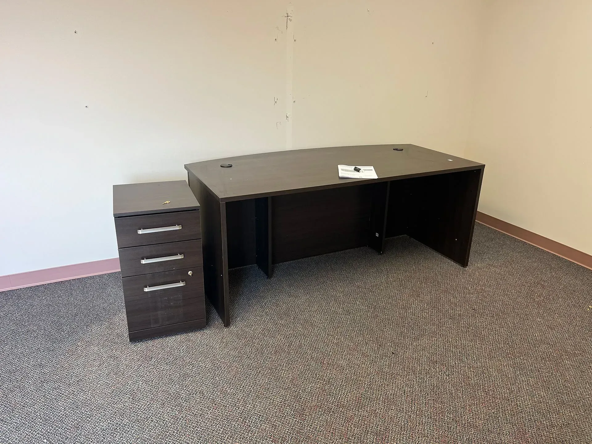 A dark brown wooden desk and a matching three-drawer filing cabinet stand in a corner on a grey carpeted floor.
