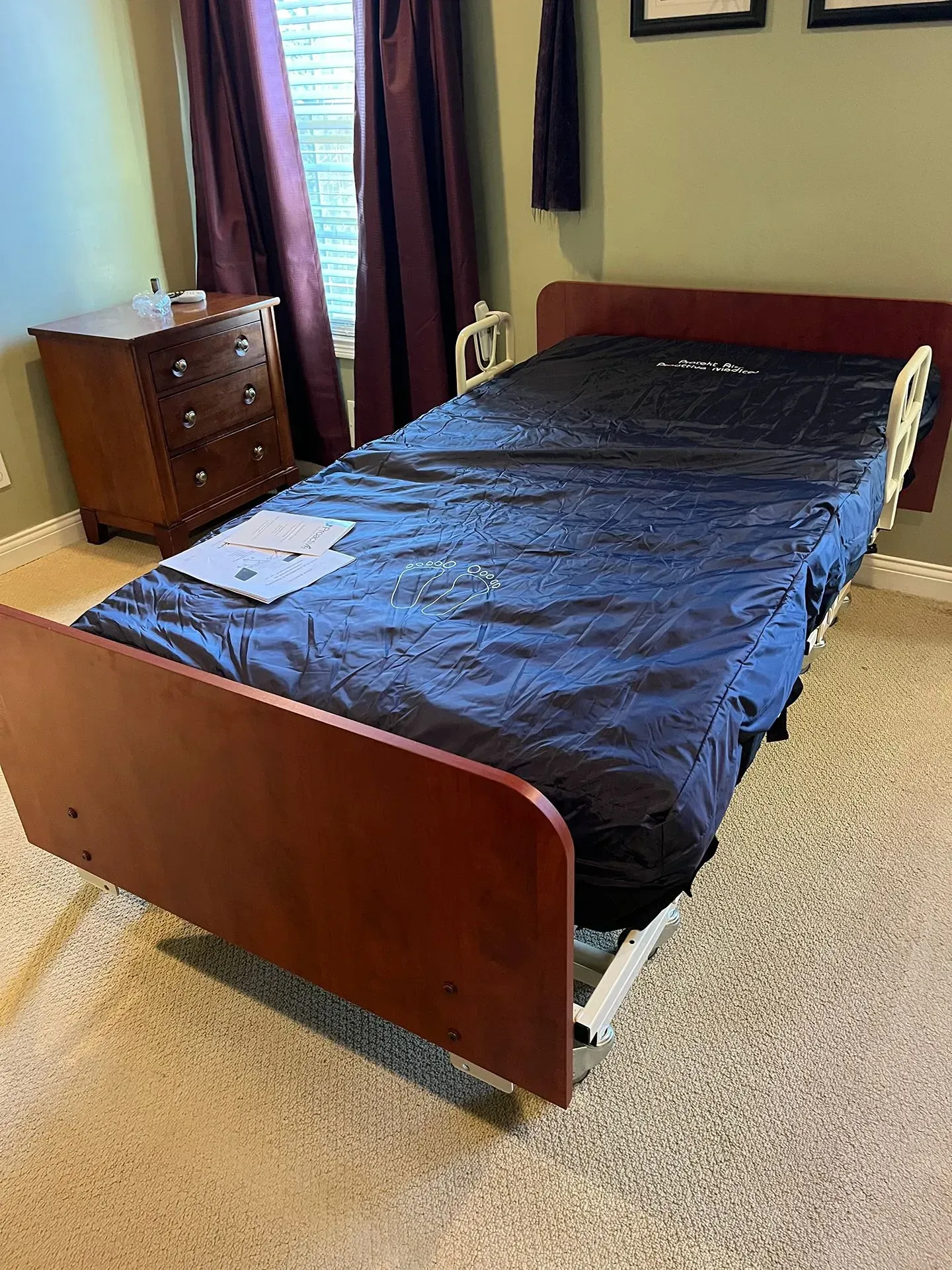 An adjustable hospital bed with a blue cover, positioned near a wooden dresser and dark curtains in a bedroom.