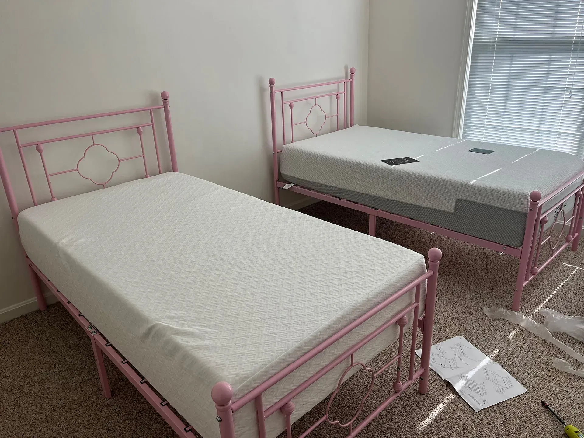 Two identical pink metal twin bed frames with mattresses, one fully assembled and one partially, on a carpeted floor.