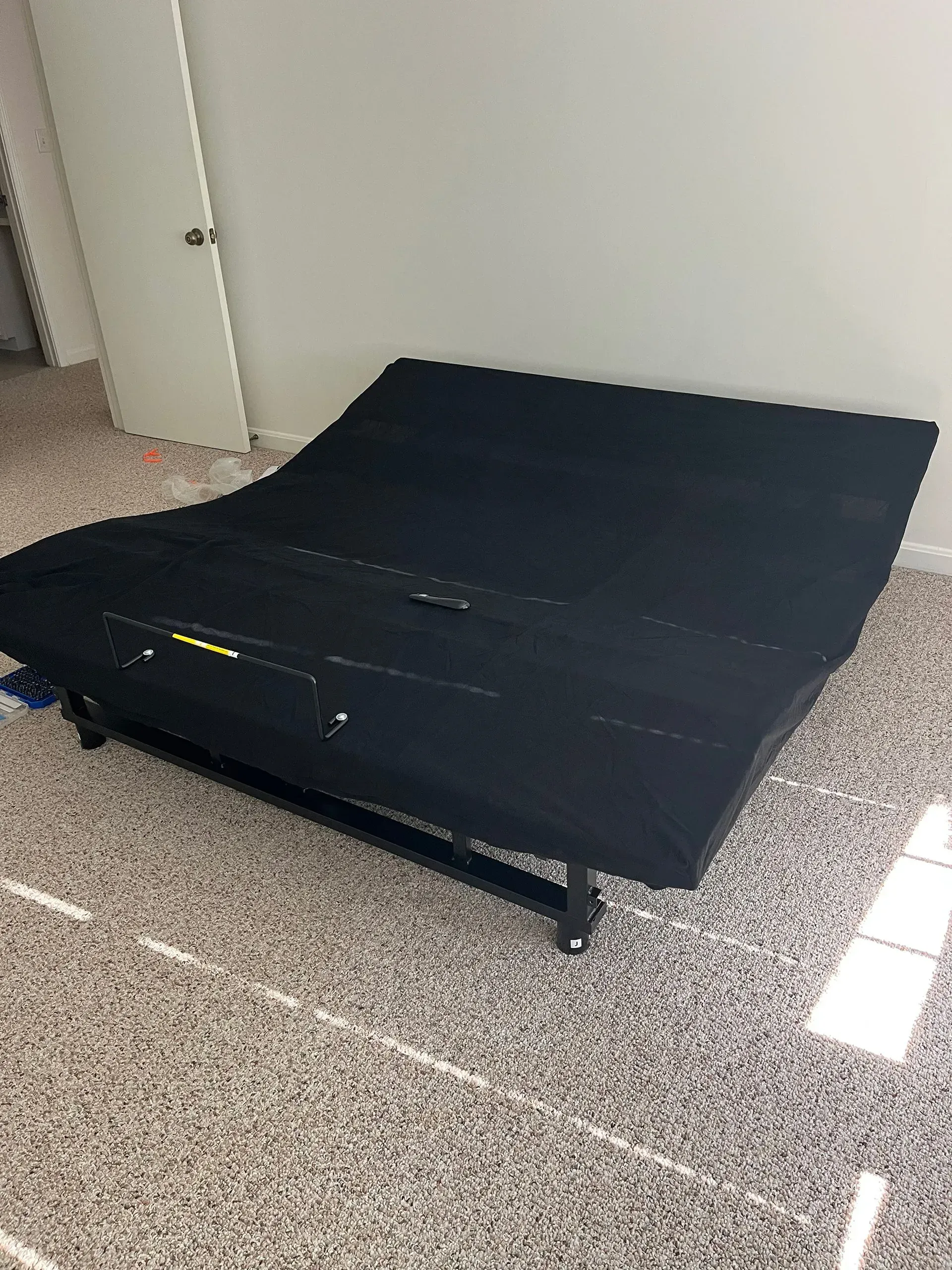 A black, adjustable bed base with a remote control positioned on top, set in an empty room with speckled flooring.