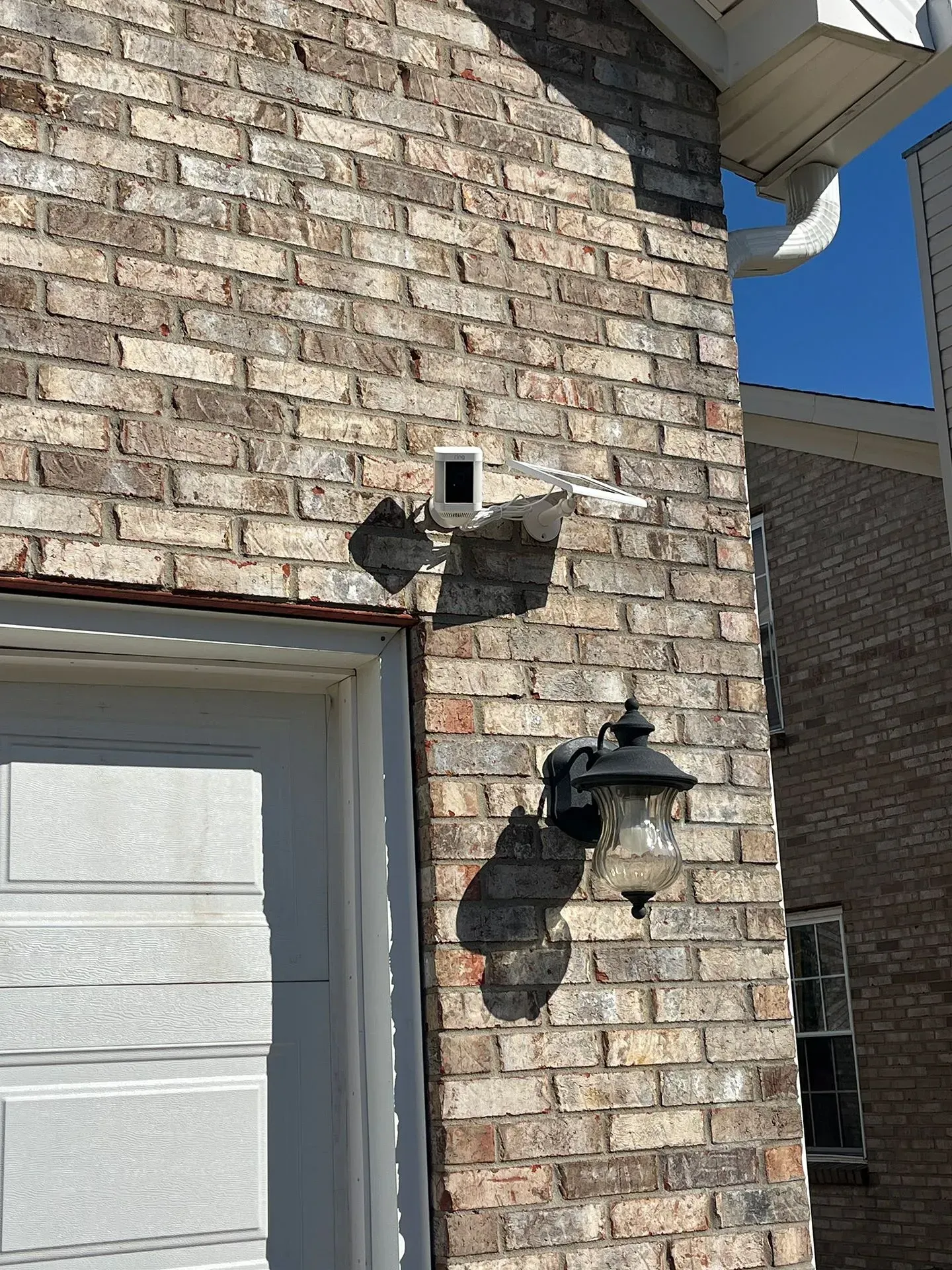 A security camera mounted on a tan brick wall above an outdoor light fixture next to a garage door.