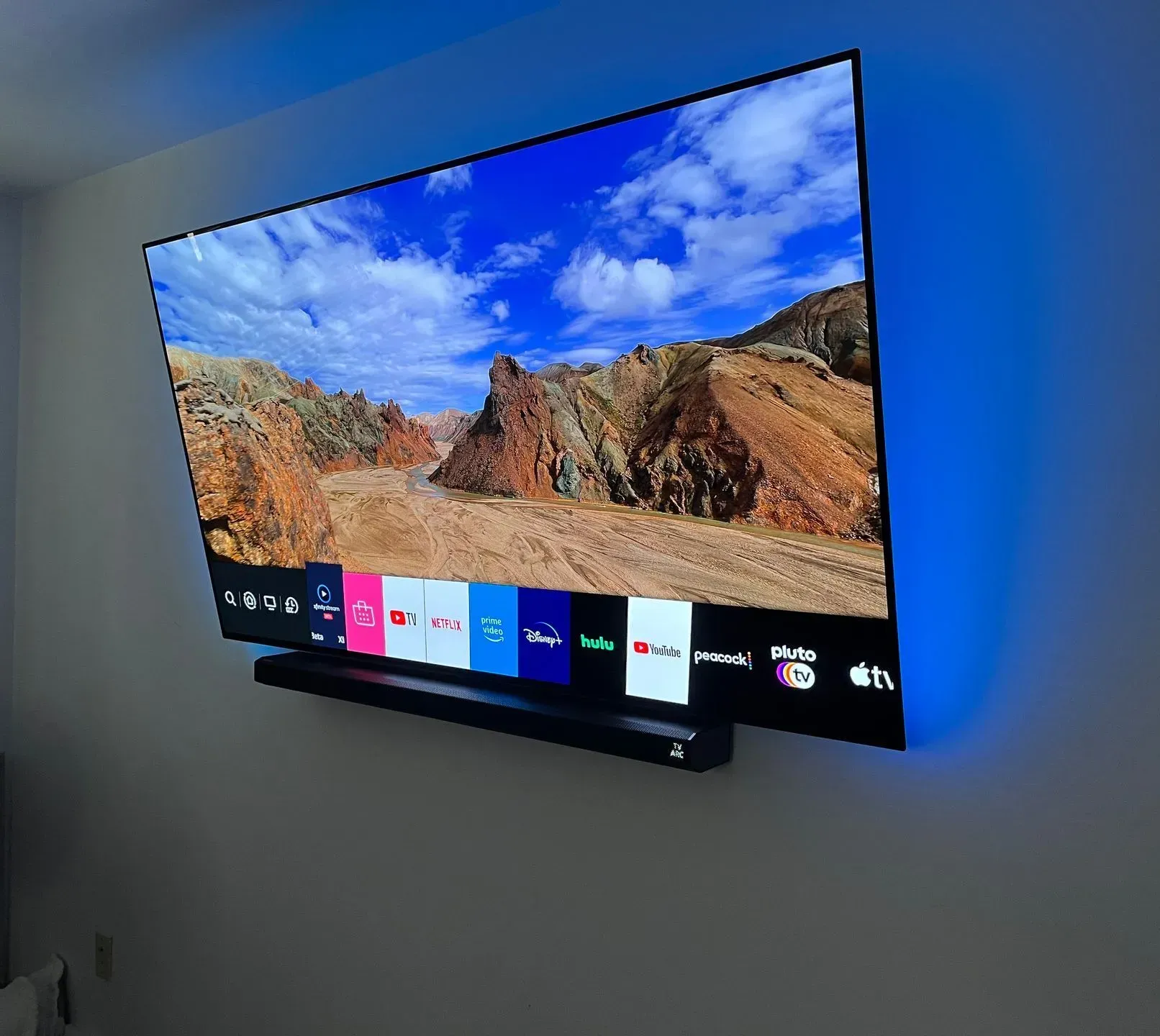 A flat-screen TV mounted on a wall with blue LED backlighting and a soundbar below, displaying a landscape background.
