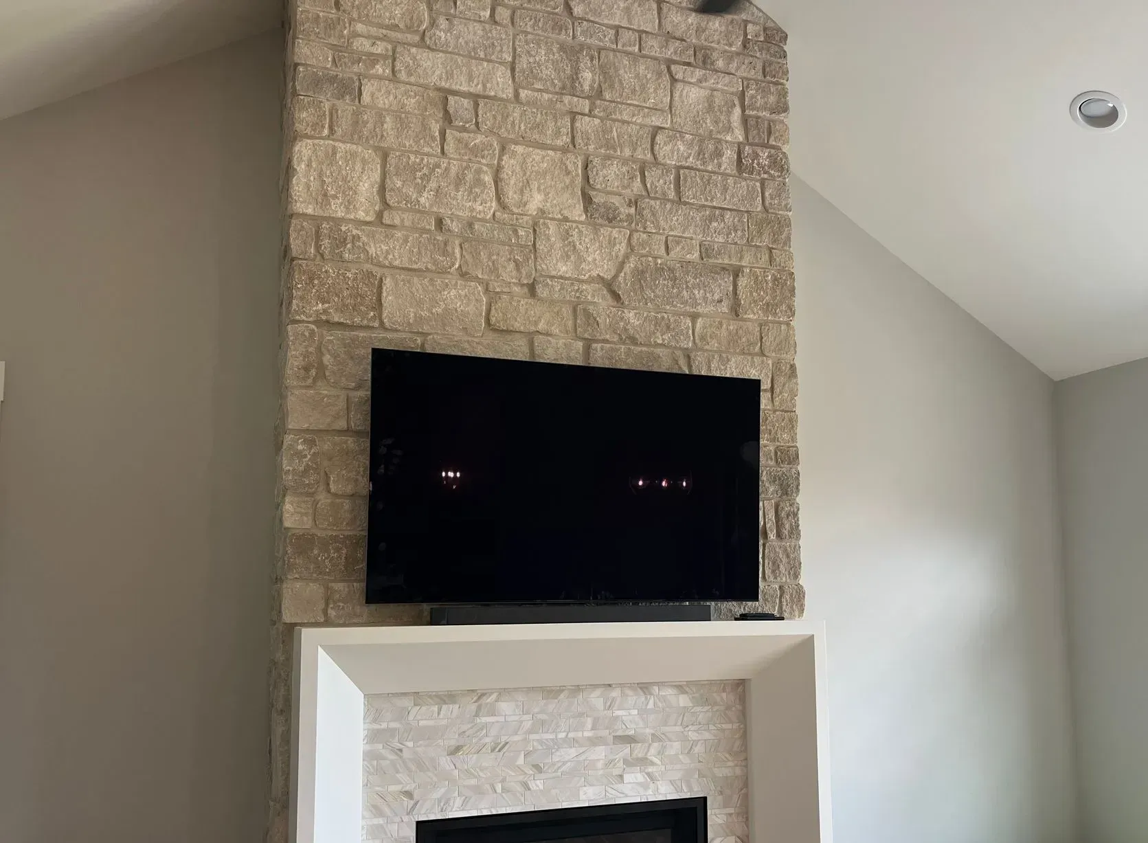 A wall-mounted TV centered on a tall, rectangular fireplace surround featuring textured, tan-colored stone masonry.