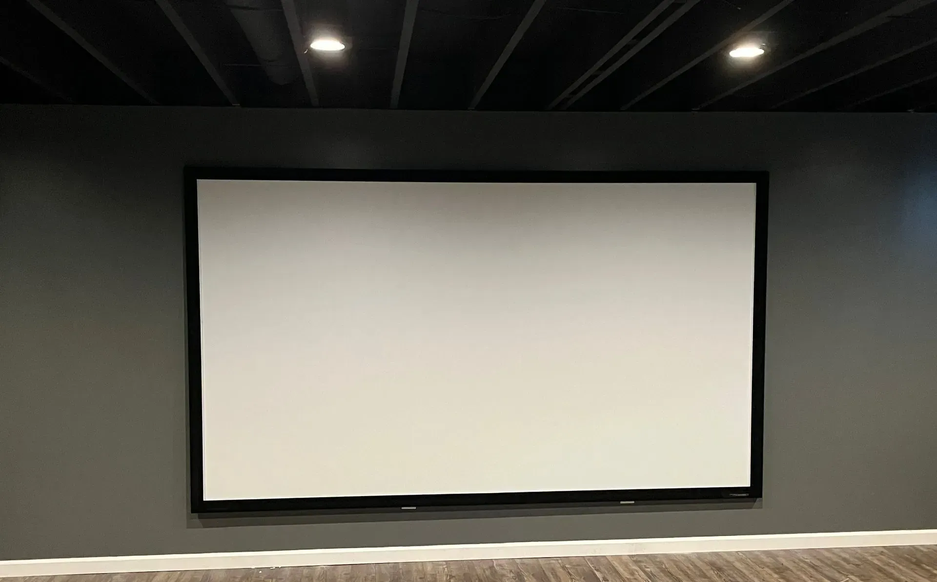 A large rectangular projector screen mounted on a dark grey wall in a room with a black, open-joist ceiling.