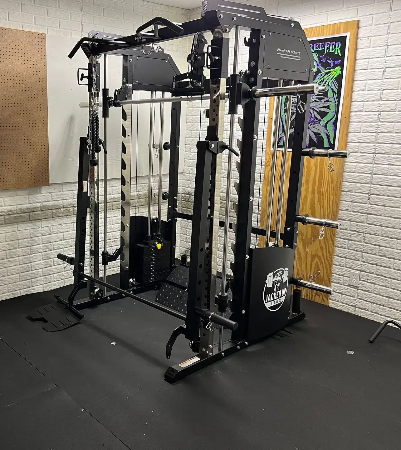 A black functional trainer and Smith machine home gym setup on black rubber flooring against a white brick wall.