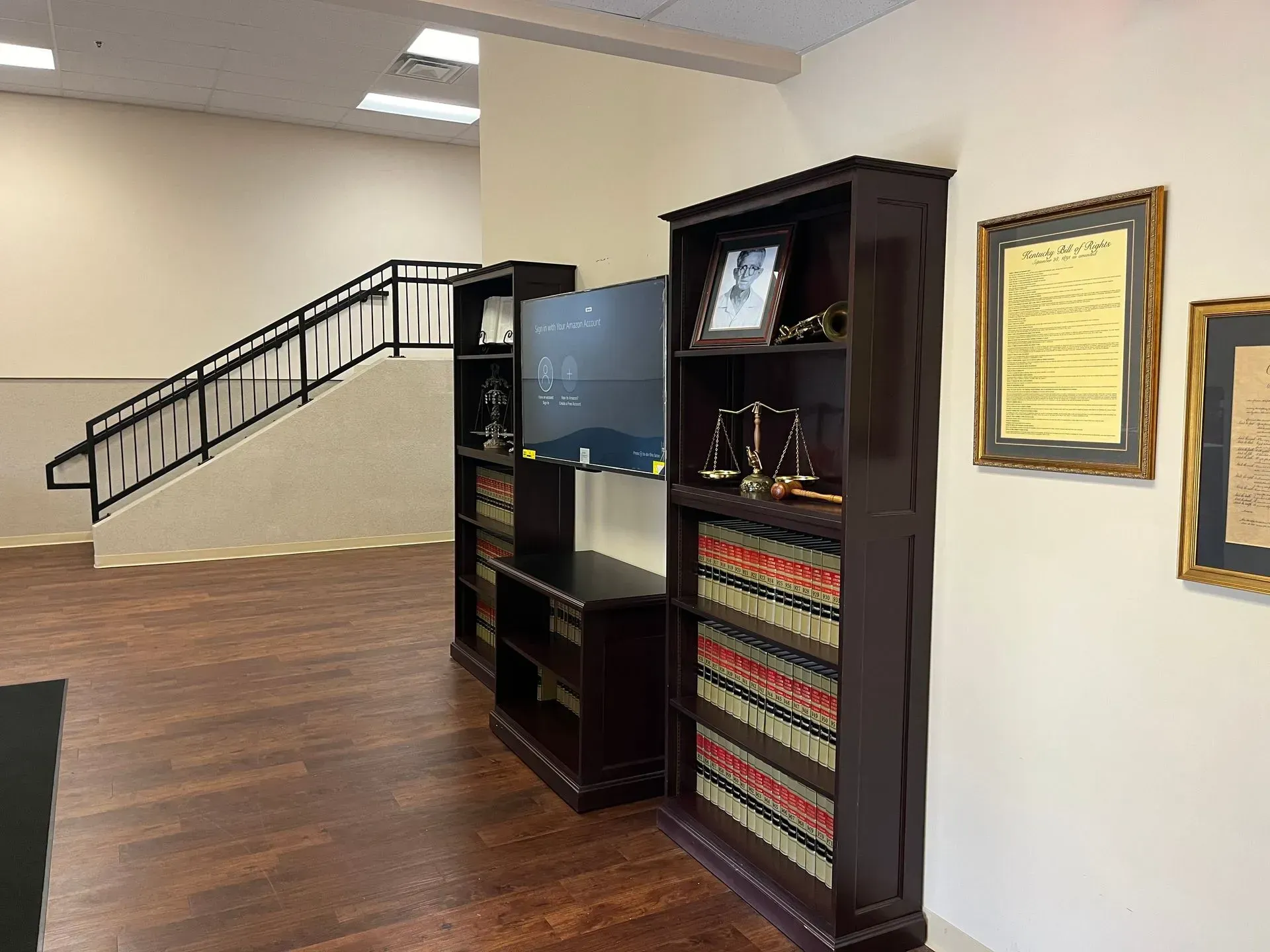 A legal office interior featuring bookshelves filled with law books, a wall-mounted monitor, and framed documents.