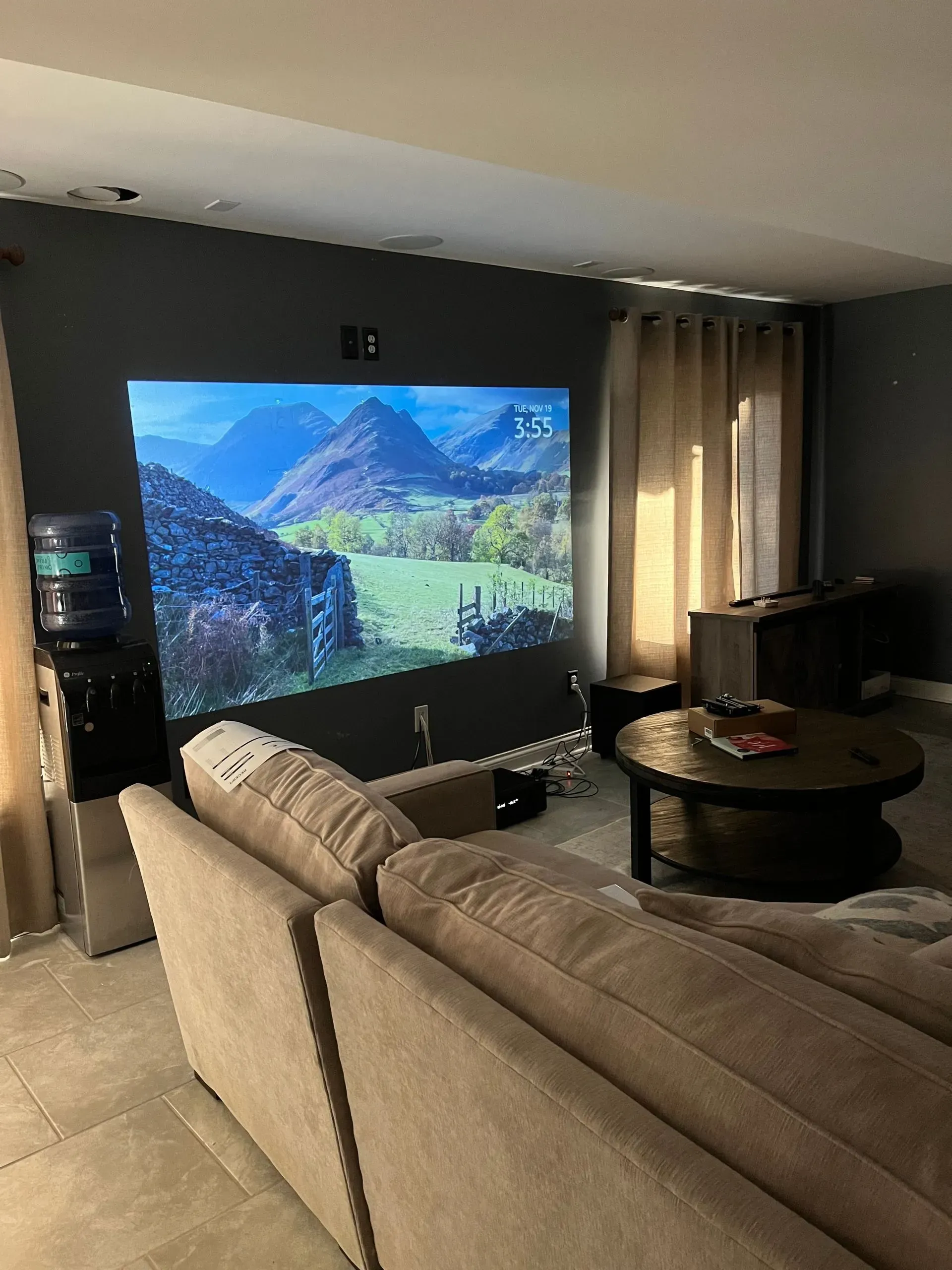 A cozy living room featuring a large projector screen showing a mountain landscape, a beige sofa, and a round coffee table.