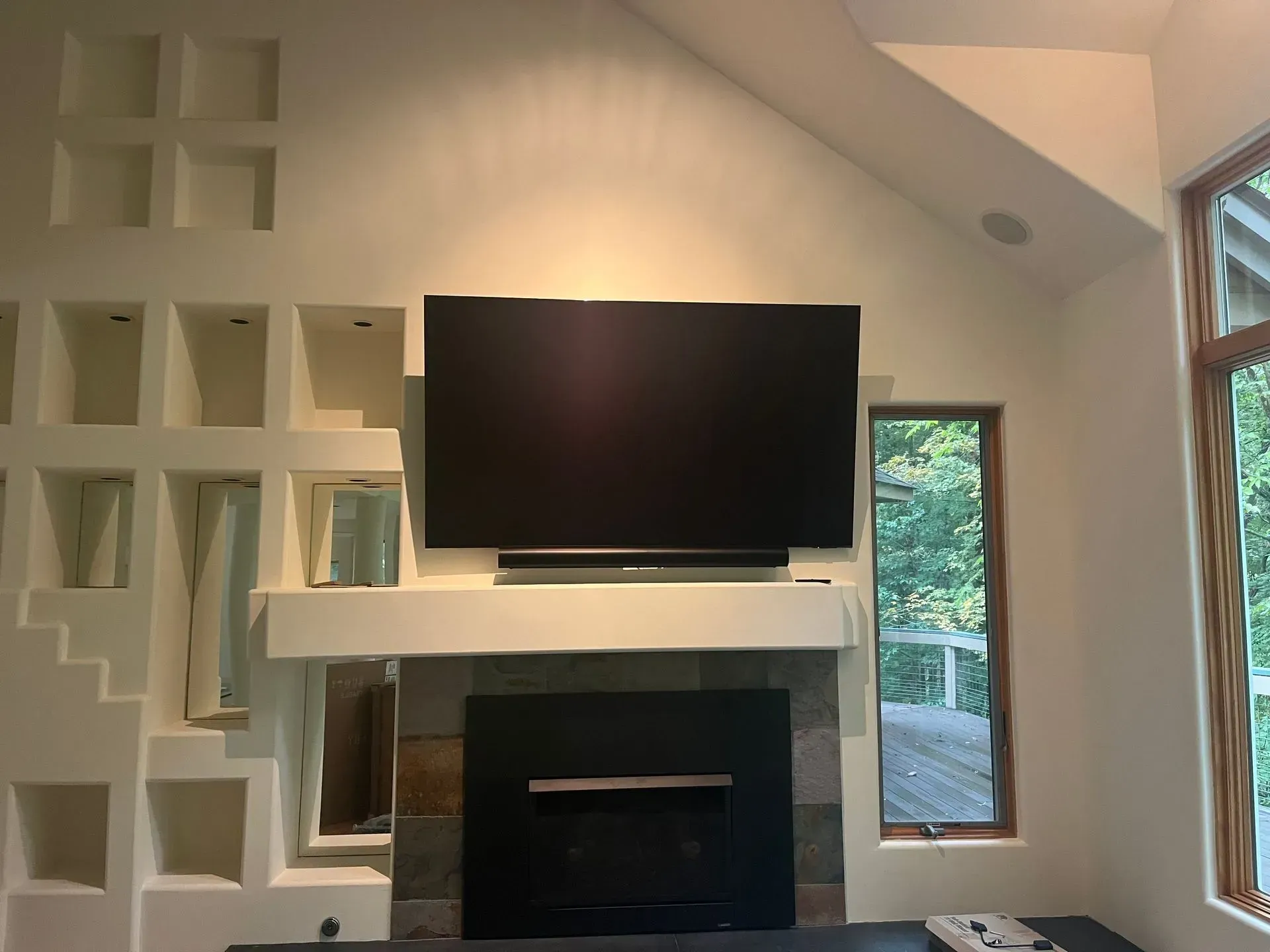 A wall-mounted TV above a fireplace, flanked by a grid of built-in wall niches and a tall window overlooking trees.