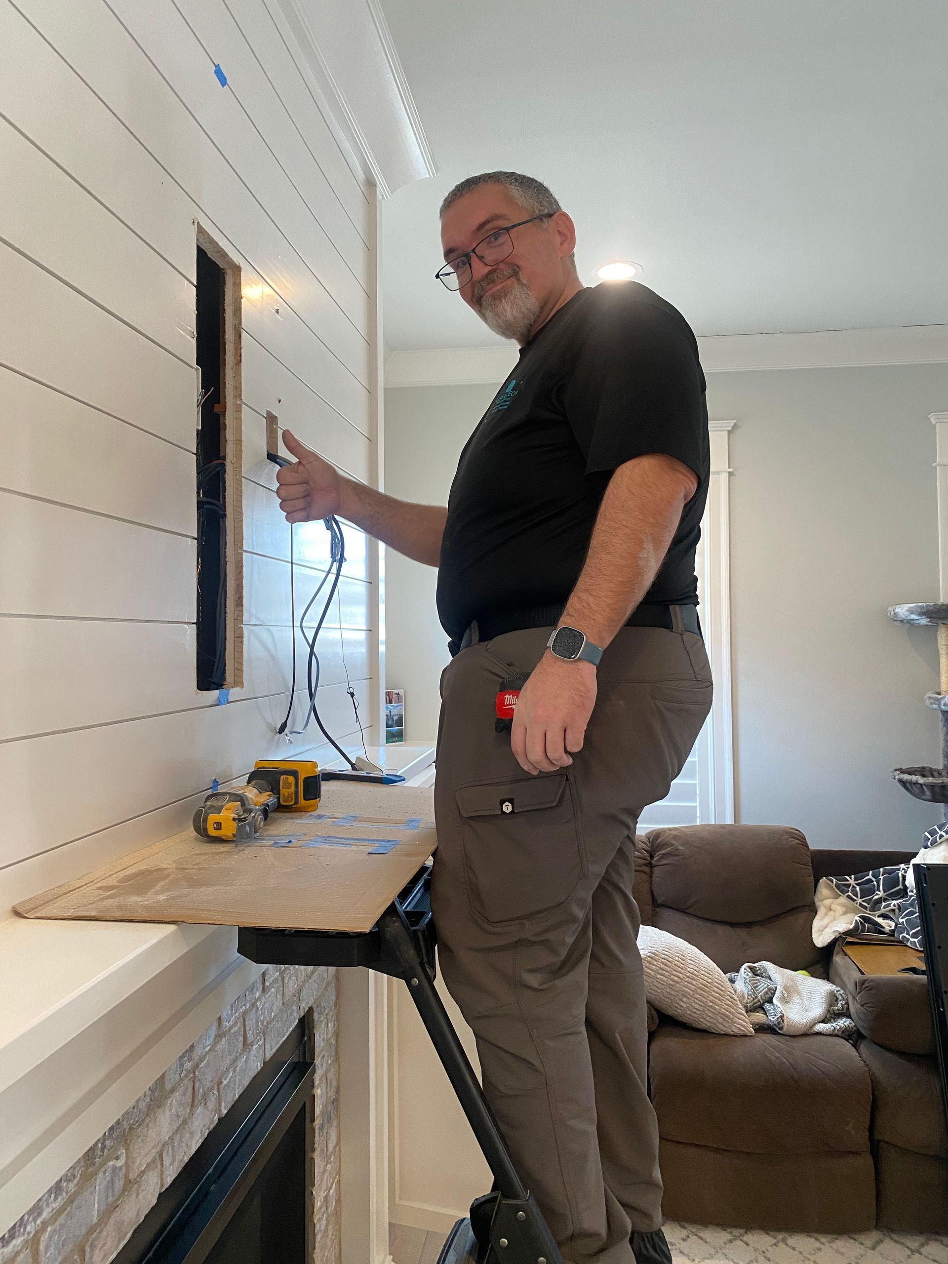 A person stands on a ladder working on a white shiplap wall, pointing at an open electrical outlet space above a fireplace.